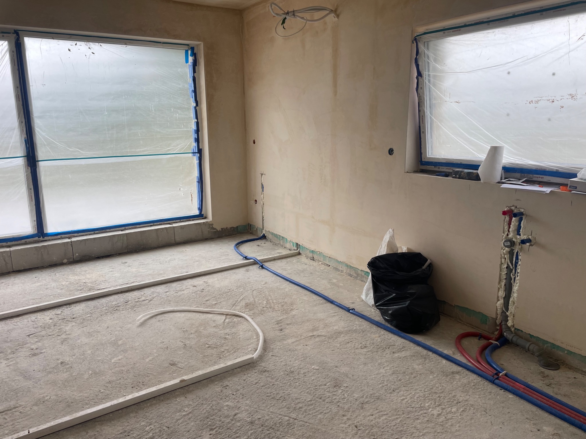 Wnętrze w trakcie prac instalacyjnych w Bydgoszczy, widoczne rury ogrzewania podłogowego, instalacja wodna w ścianie zabezpieczonej pianką oraz okna zakryte folią ochronną.
