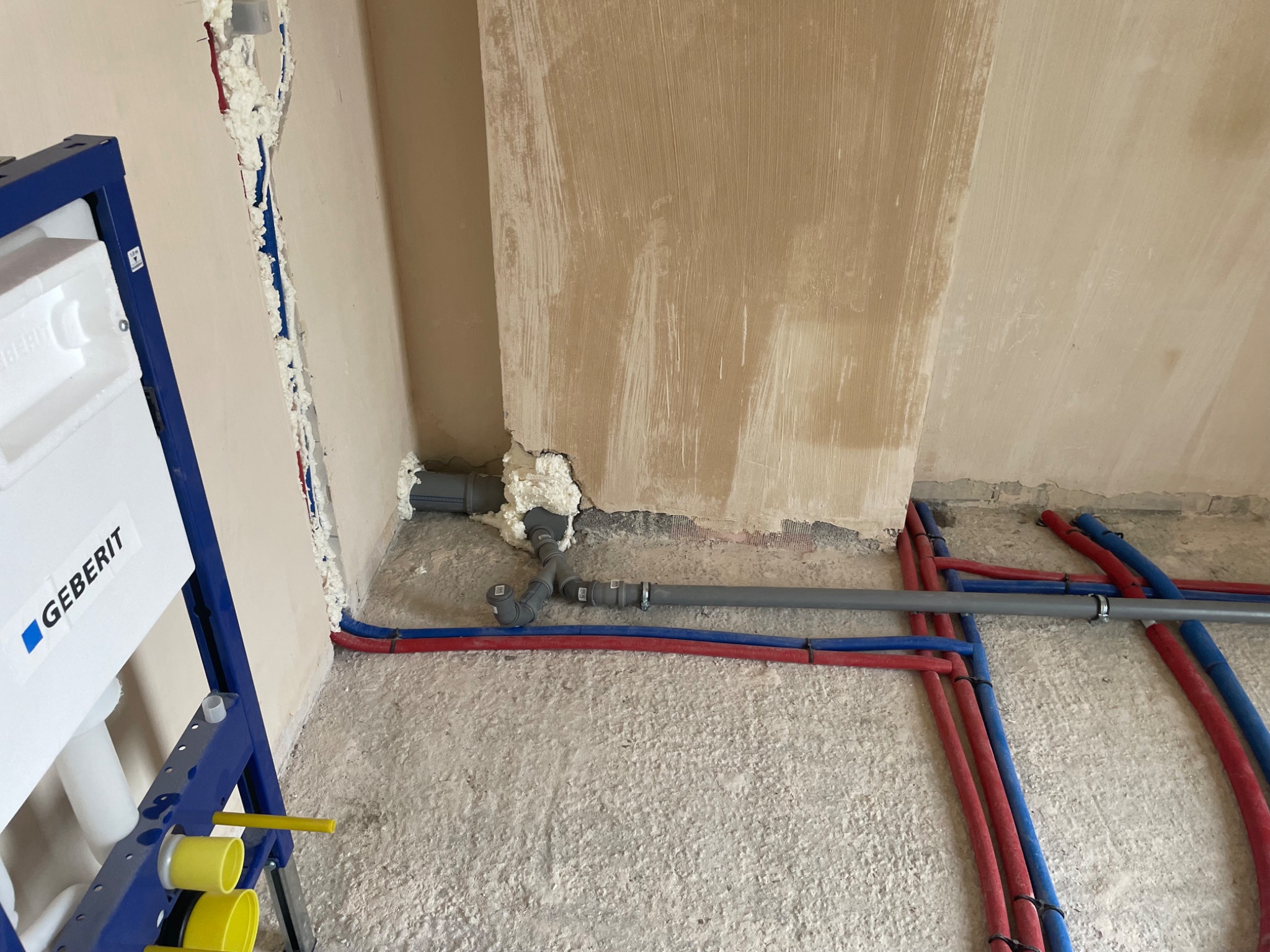 Surowe wnętrze łazienki w trakcie instalacji: widoczne rury wodne (czerwone i niebieskie), szare rury kanalizacyjne, stelaż podtynkowy Geberit oraz ślady pianki montażowej na ścianach.