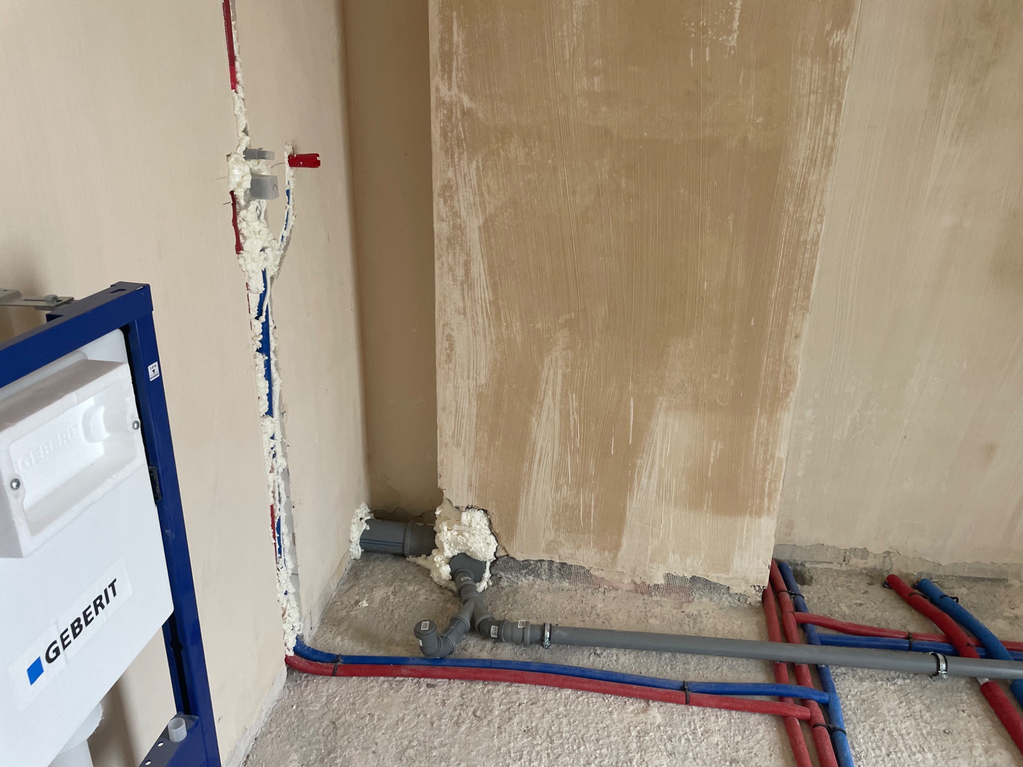 Montaż instalacji wodnej z niebieskimi i czerwonymi rurami w bruzdach ściennych, pianka montażowa uszczelniająca, stelaż podtynkowy Geberit z widocznym logo, surowy beton na podłodze i ściany...