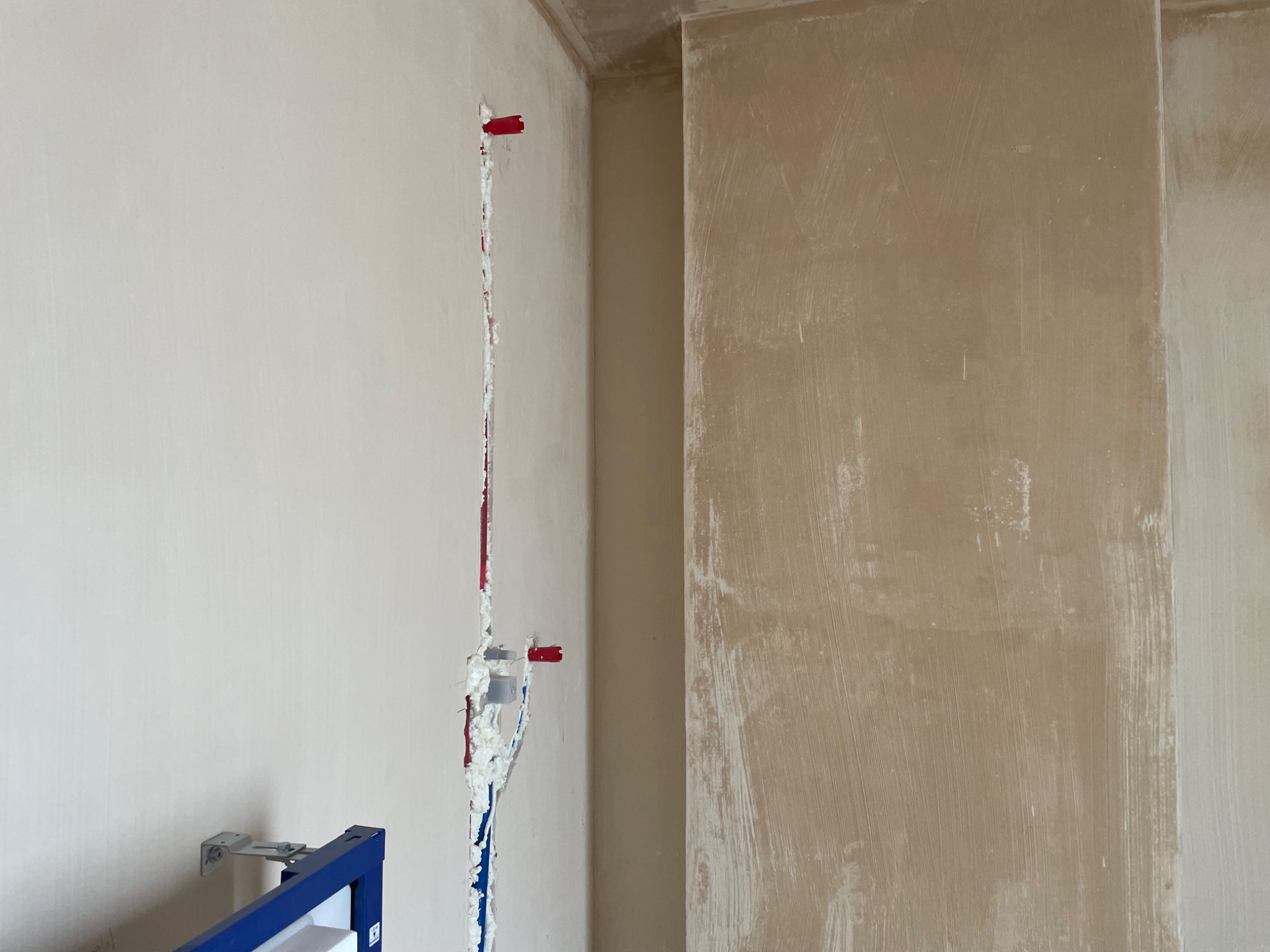Wykuta bruzda w ścianie z widocznymi rurami instalacji wodnej i pianką montażową, fragment stelaża podtynkowego WC.