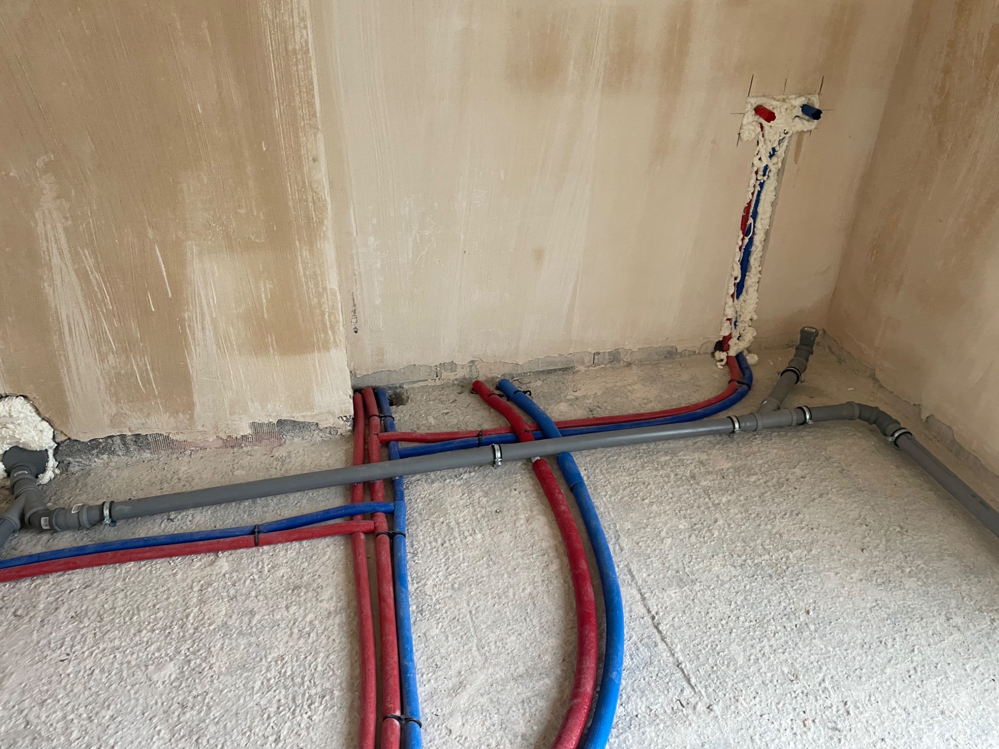 Surowe ściany w trakcie remontu z widocznymi rurami instalacji wodnej w kolorze czerwonym i niebieskim, oraz szarymi rurami kanalizacyjnymi, częściowo zabezpieczonymi pianką montażową.