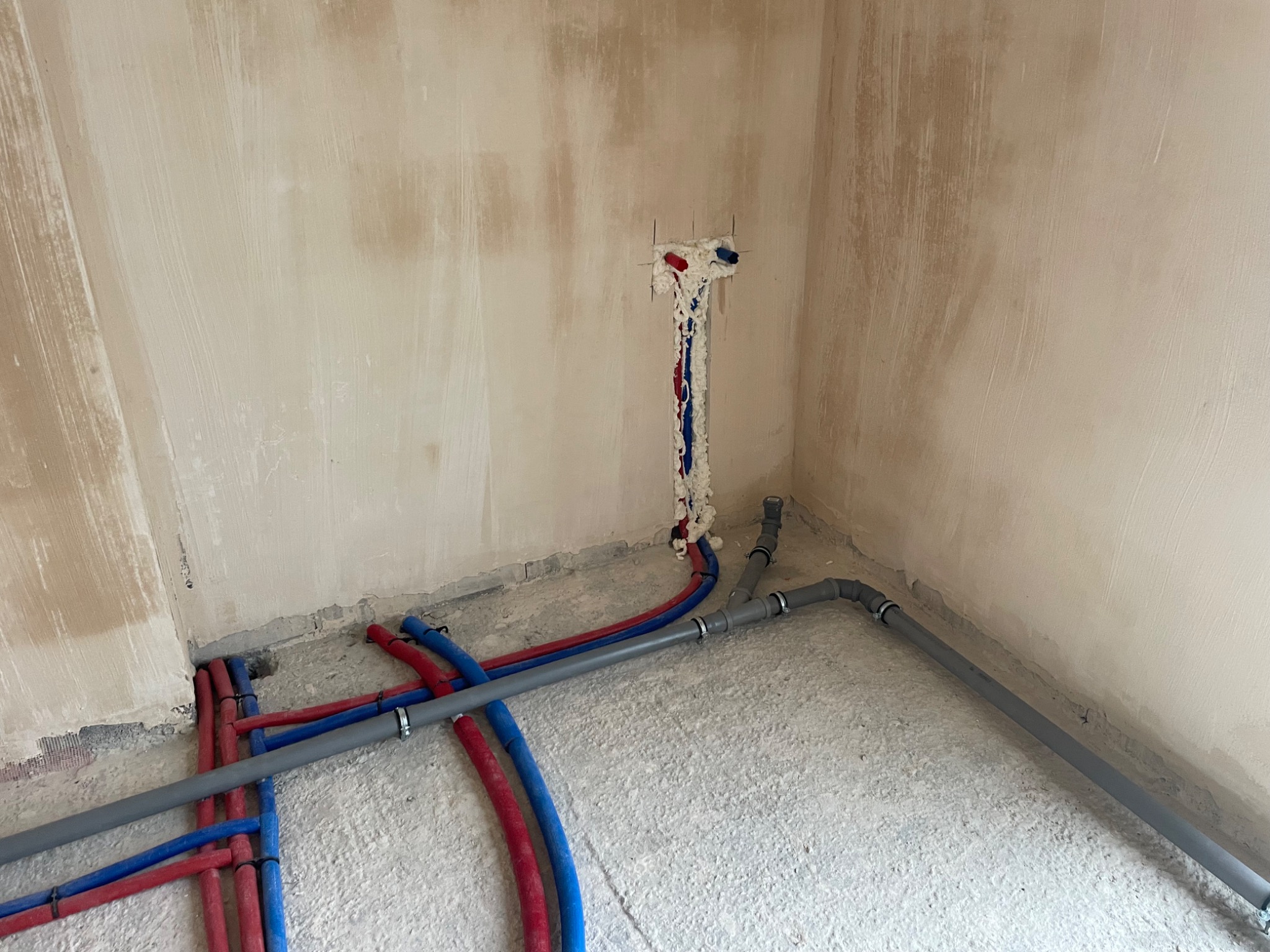 Surowe wnętrze w trakcie prac hydraulicznych: widoczne rury doprowadzające wodę (czerwone i niebieskie) wystające ze ściany, zabezpieczone pianką montażową, oraz szare rury kanalizacyjne prowadzone...