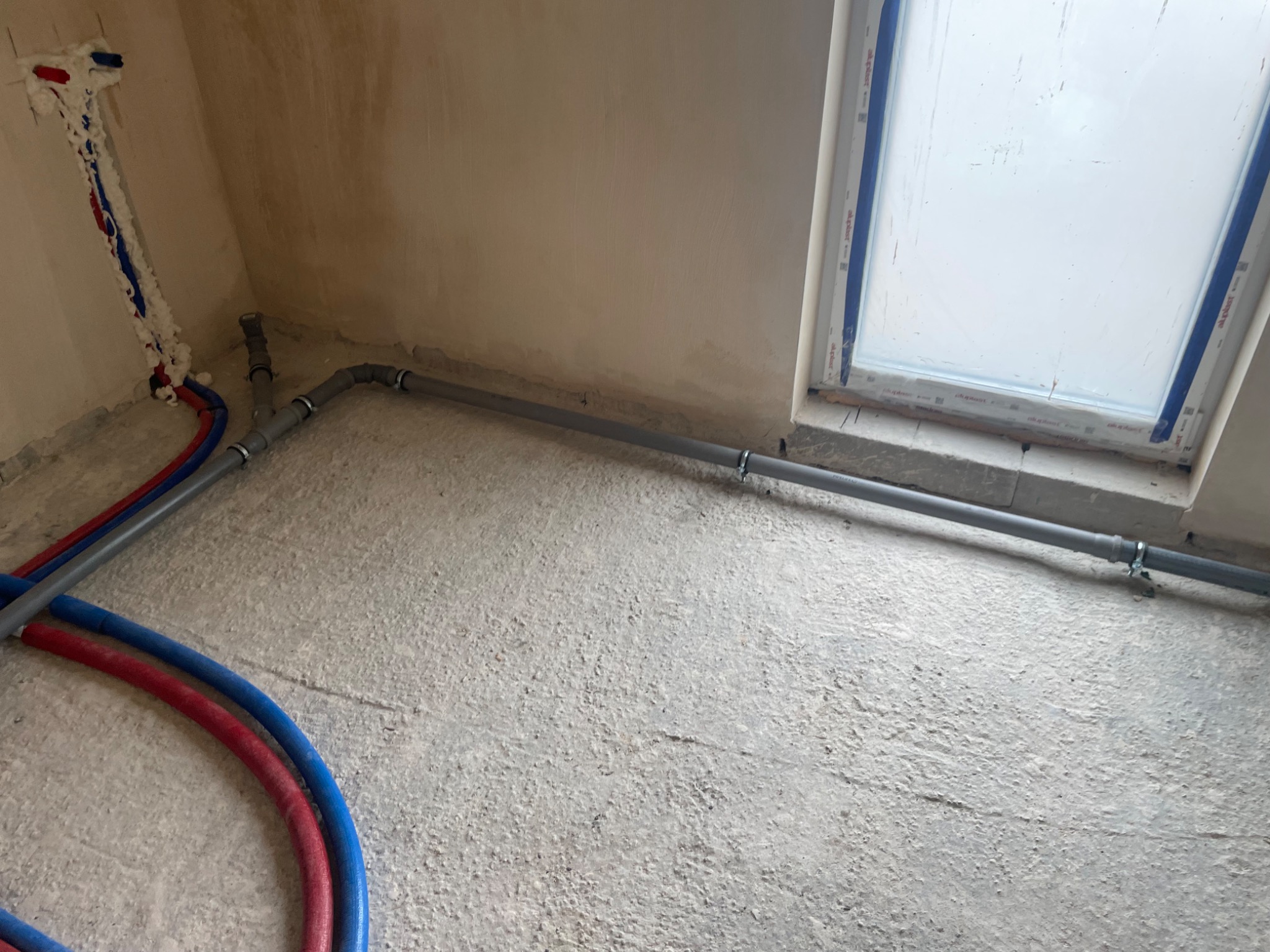 Szare rury kanalizacyjne i czerwono-niebieskie rury instalacji wodnej ułożone na surowym, betonowym podłożu w trakcie prac instalacyjnych, widoczne fragmenty ścian i okno dachowe.