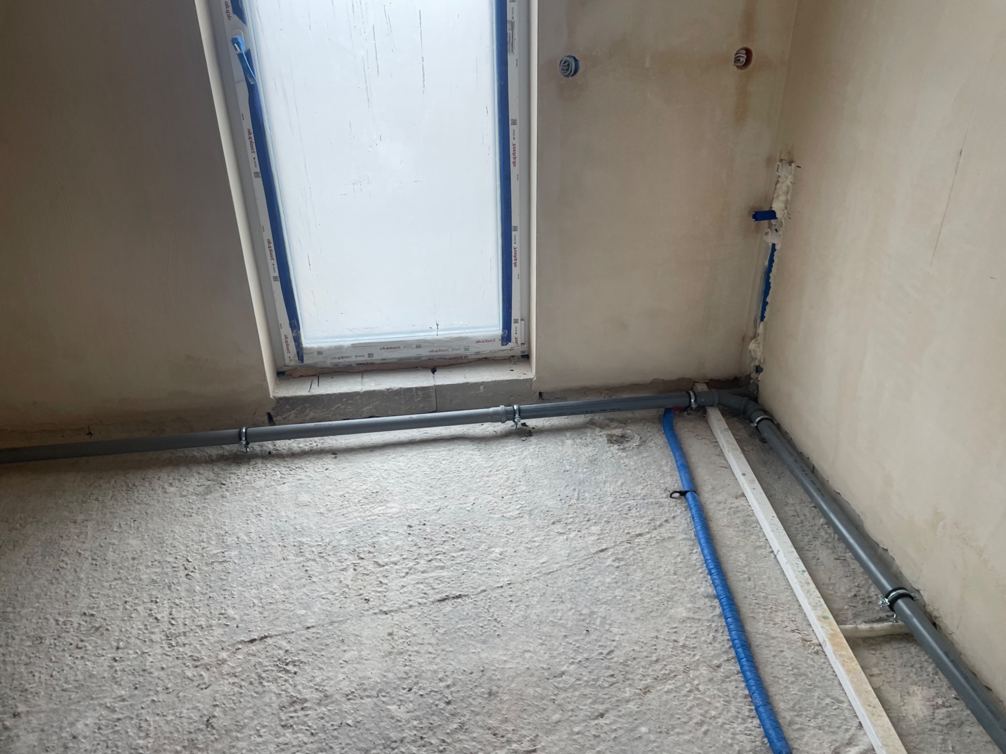 Szare i niebieskie rury instalacyjne prowadzone wzdłuż ściany i pod oknem dachowym w surowym wnętrzu, widoczne mocowania do ściany i podłoża.