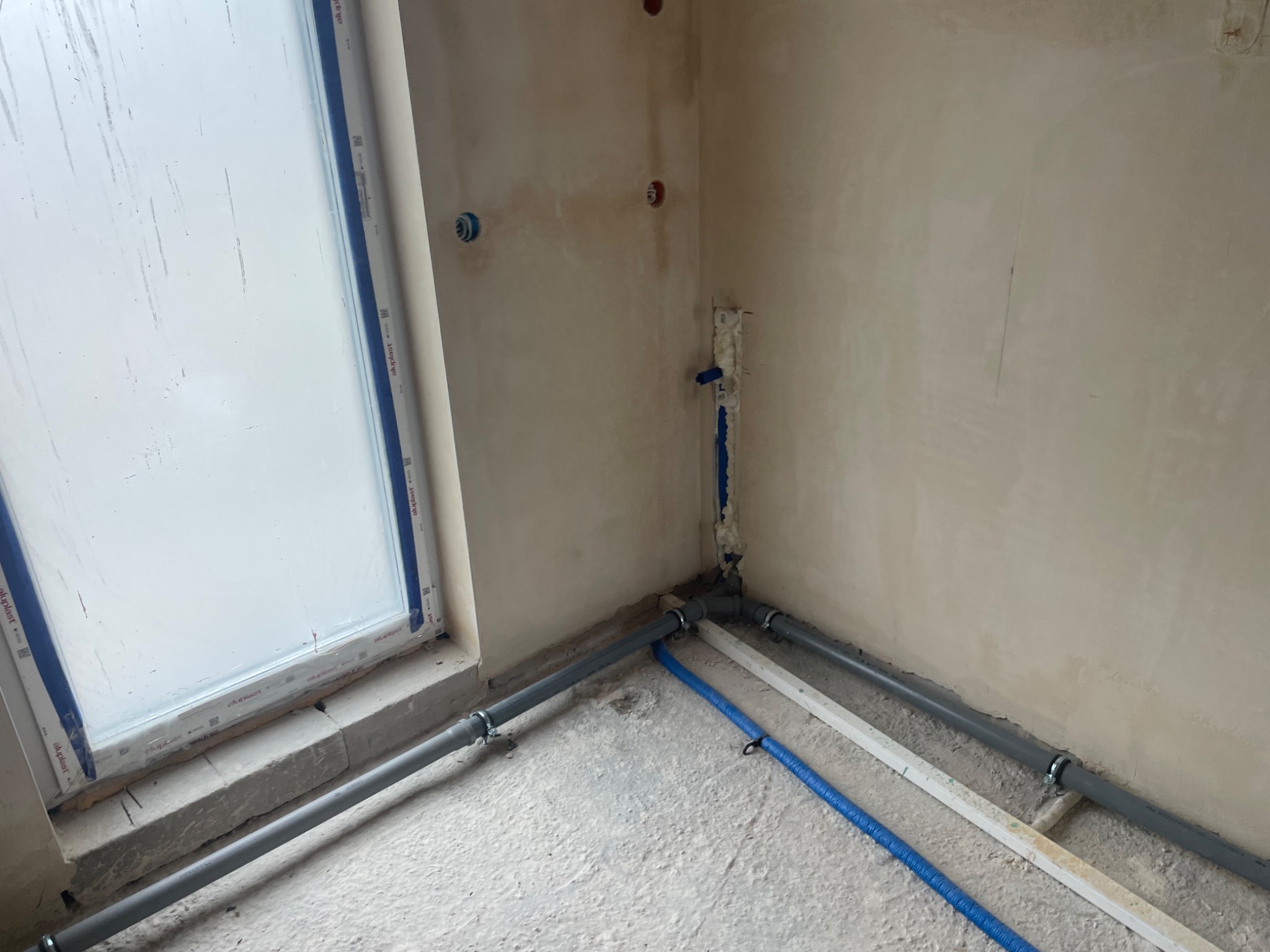 Szare i niebieskie rury instalacyjne ułożone na podłodze w surowym wnętrzu w Bydgoszczy, widoczne przyłącza do ściany i okna.