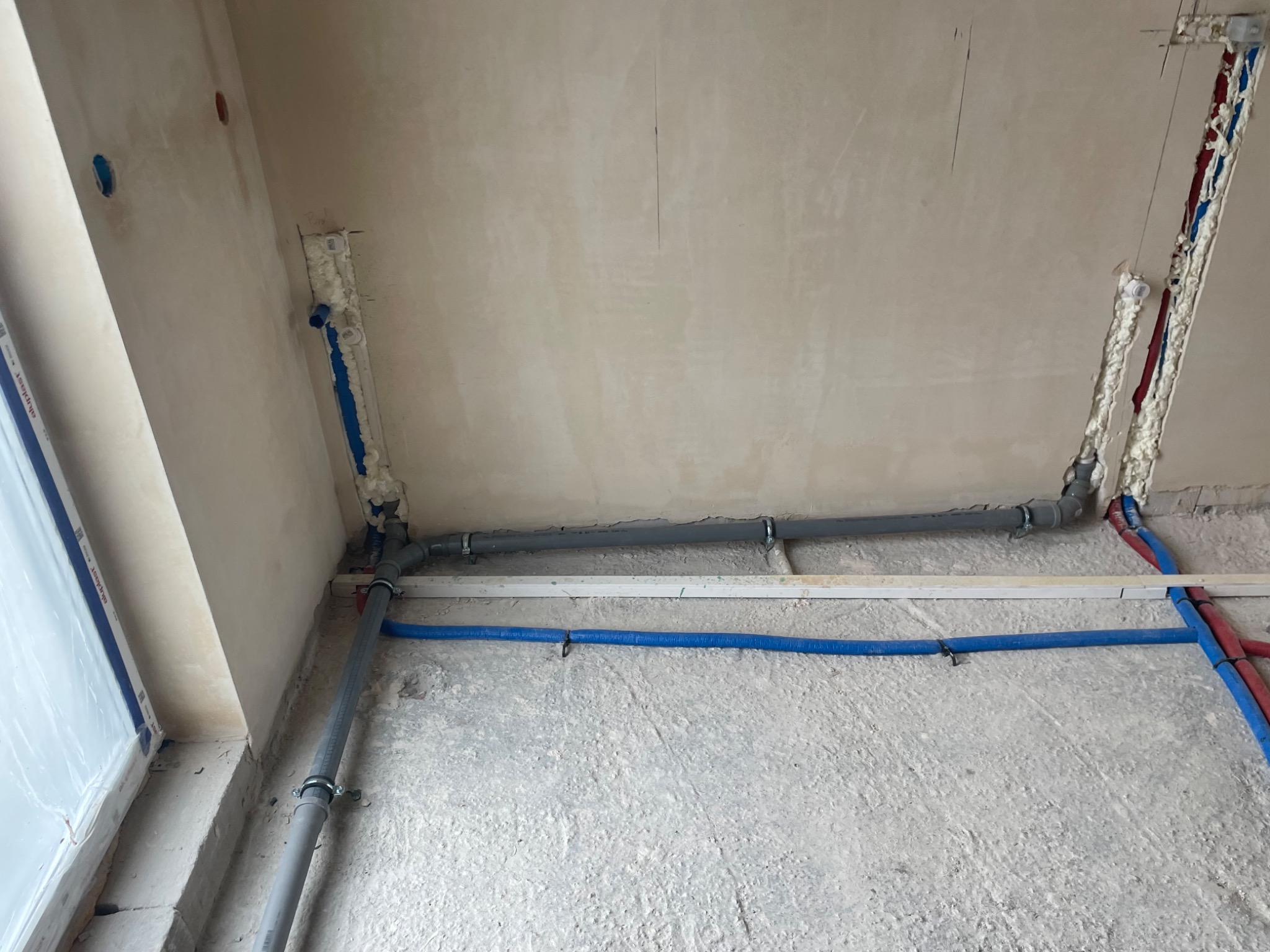 Surowe wnętrze w trakcie prac instalacyjnych: szare rury kanalizacyjne z obejmami, niebieskie i czerwone rury w izolacji piankowej w ścianie, surowy beton na podłodze, widoczne otwory w ścianie.