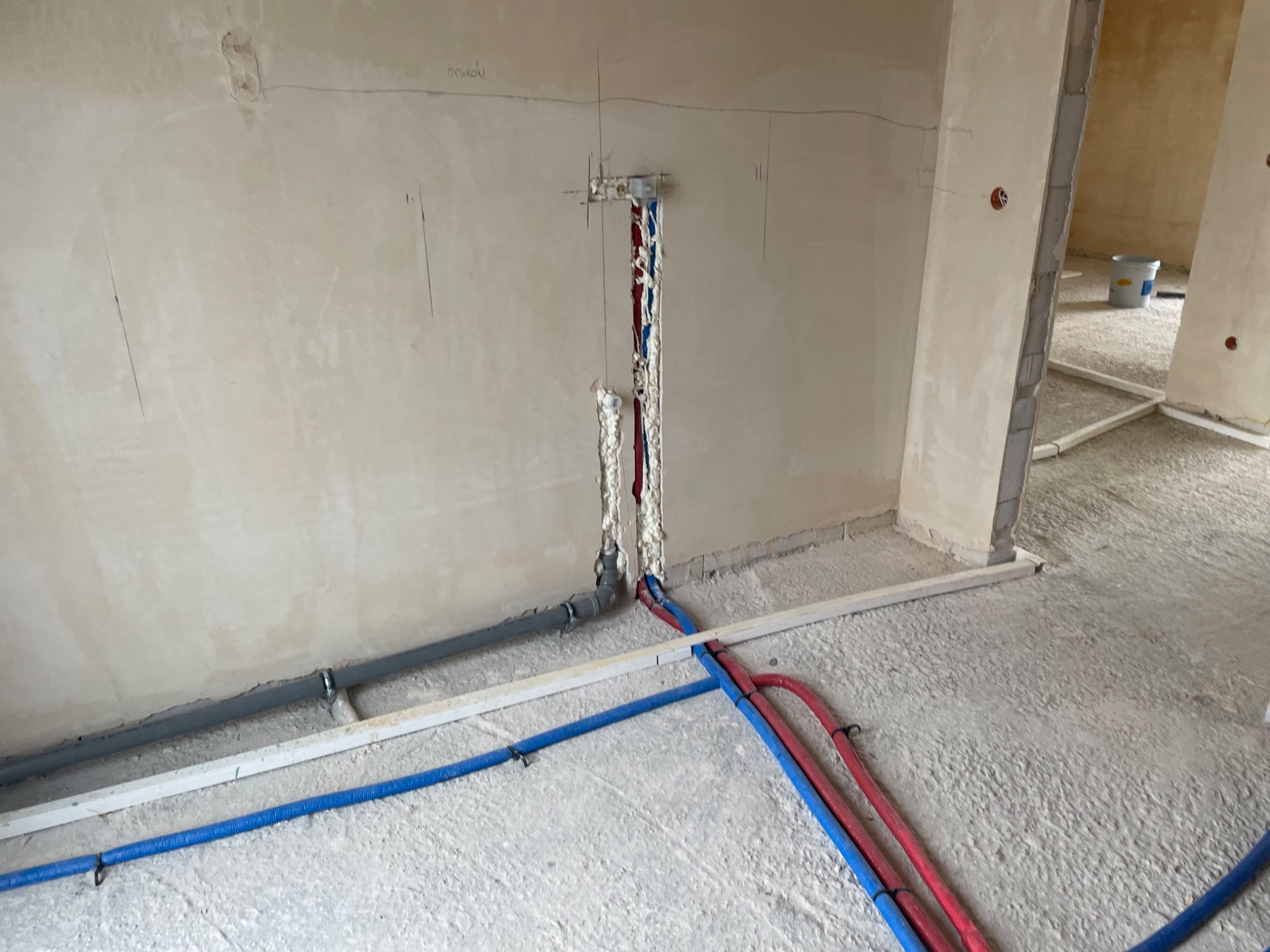 Surowy stan instalacji hydraulicznej w nowym budynku: wystające rury PEX (czerwone i niebieskie) oraz szare rury kanalizacyjne, zabezpieczone pianką montażową w ścianie, widoczne oznaczenia...