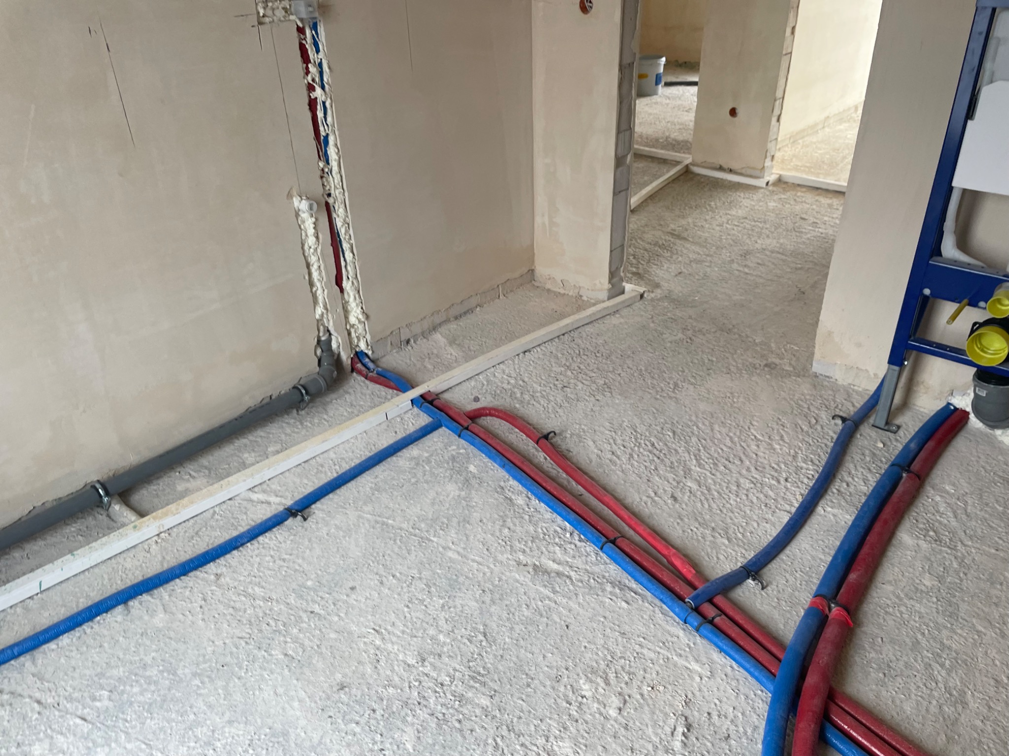 Surowe wnętrze w trakcie remontu z widocznymi nieotynkowanymi ścianami, podłogą w trakcie wylewki i rozłożonymi rurami instalacji wodnej i grzewczej w kolorach niebieskim i czerwonym...