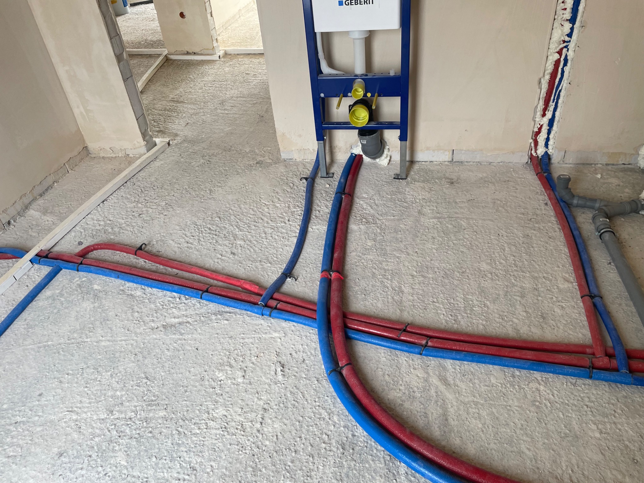 Nowa instalacja wodna z rurami PEX w kolorze czerwonym i niebieskim, zamocowanymi do podłoża w surowym stanie, z widoczną podtynkową spłuczką Geberit.