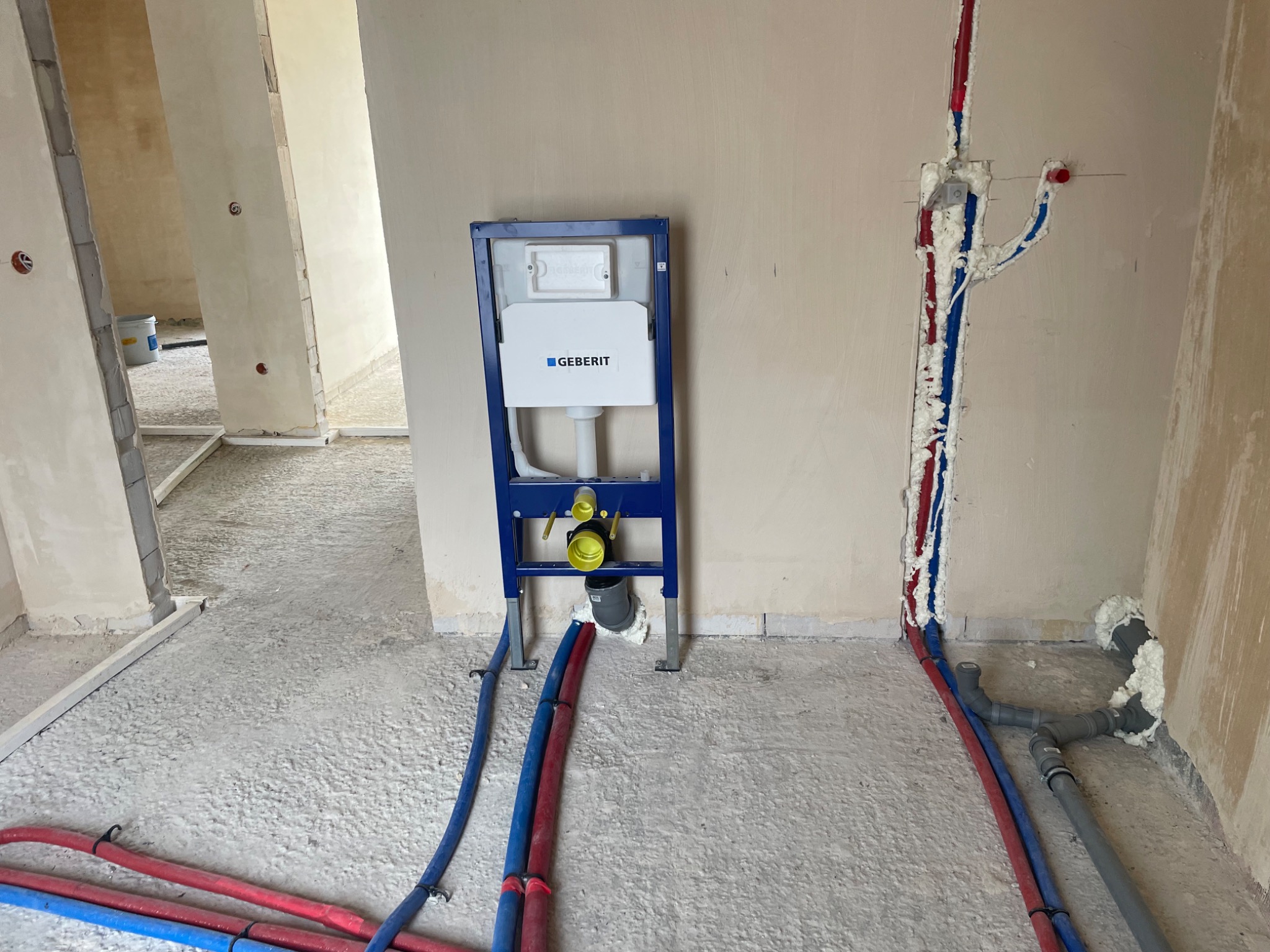 Stelaż podtynkowy Geberit zamontowany w surowym wnętrzu łazienki, widoczne rury instalacji wodnej i kanalizacyjnej zabezpieczone pianką montażową.