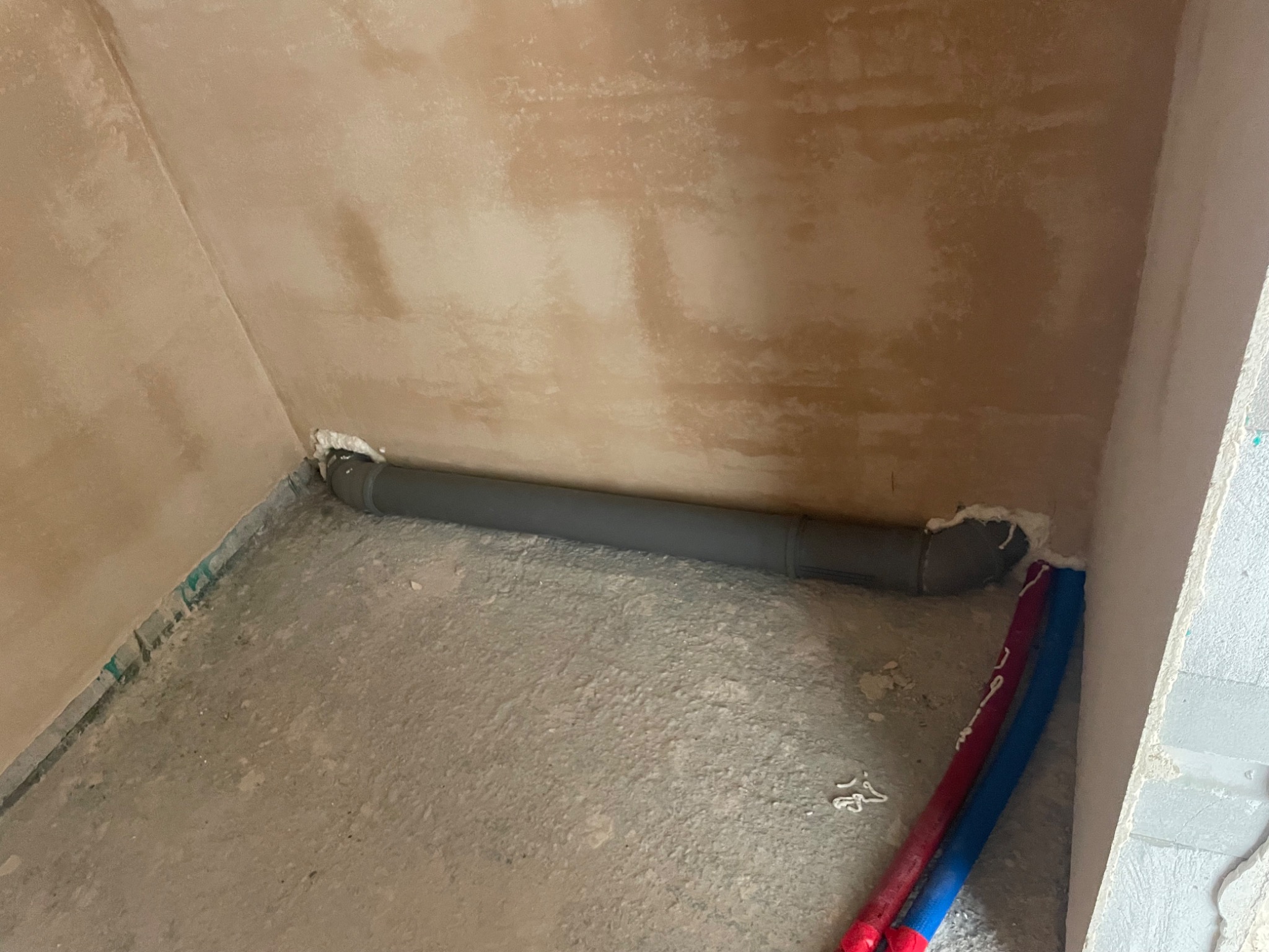 Szara rura kanalizacyjna poziomo przy ścianie, uszczelniona pianką montażową, obok czerwona i niebieska rura instalacji wodnej, surowy tynk na ścianach.