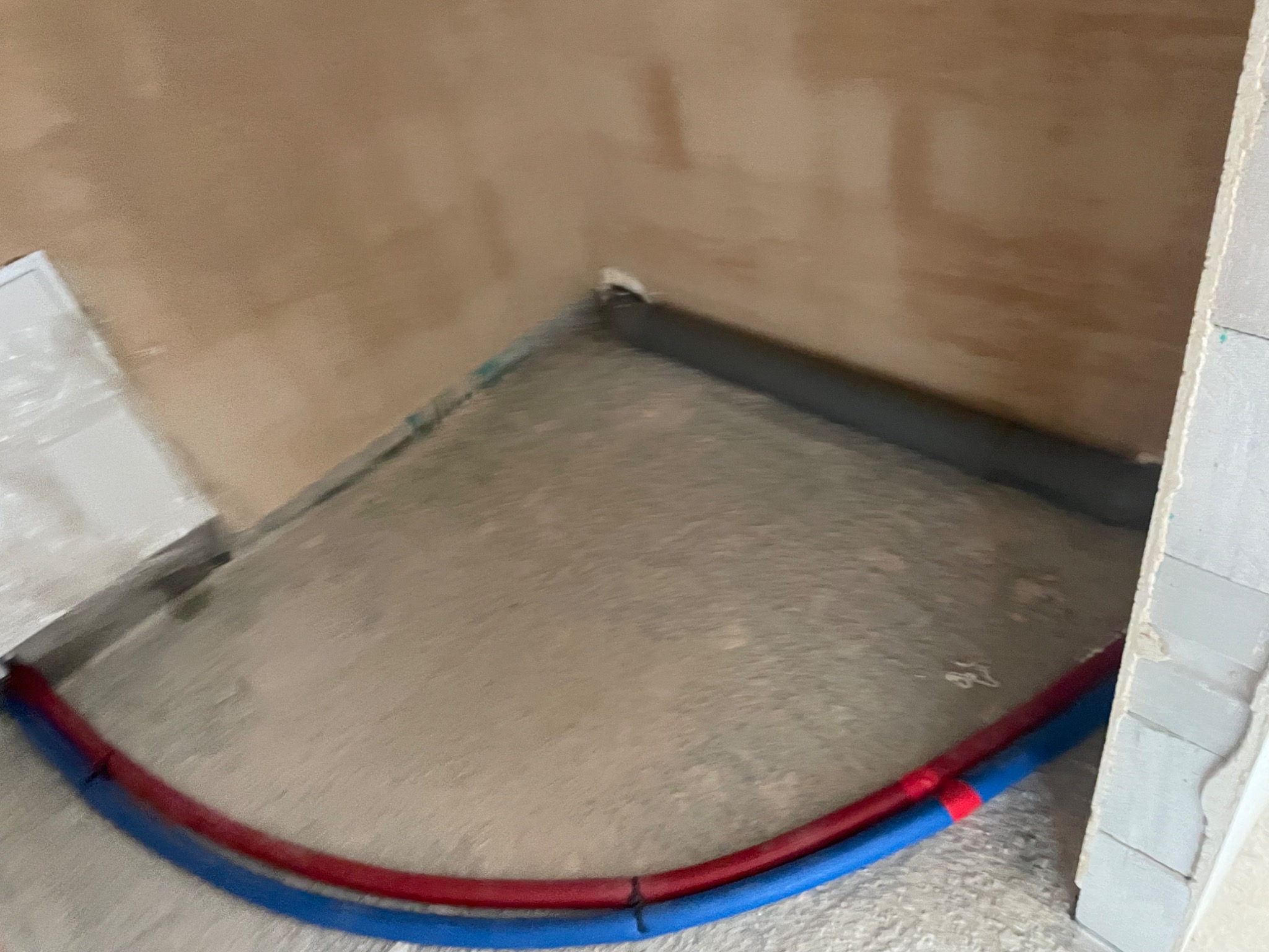 Wylewka pod ogrzewanie podłogowe z widocznymi rurami w kolorze niebieskim i czerwonym, przy ścianie z surowym tynkiem.