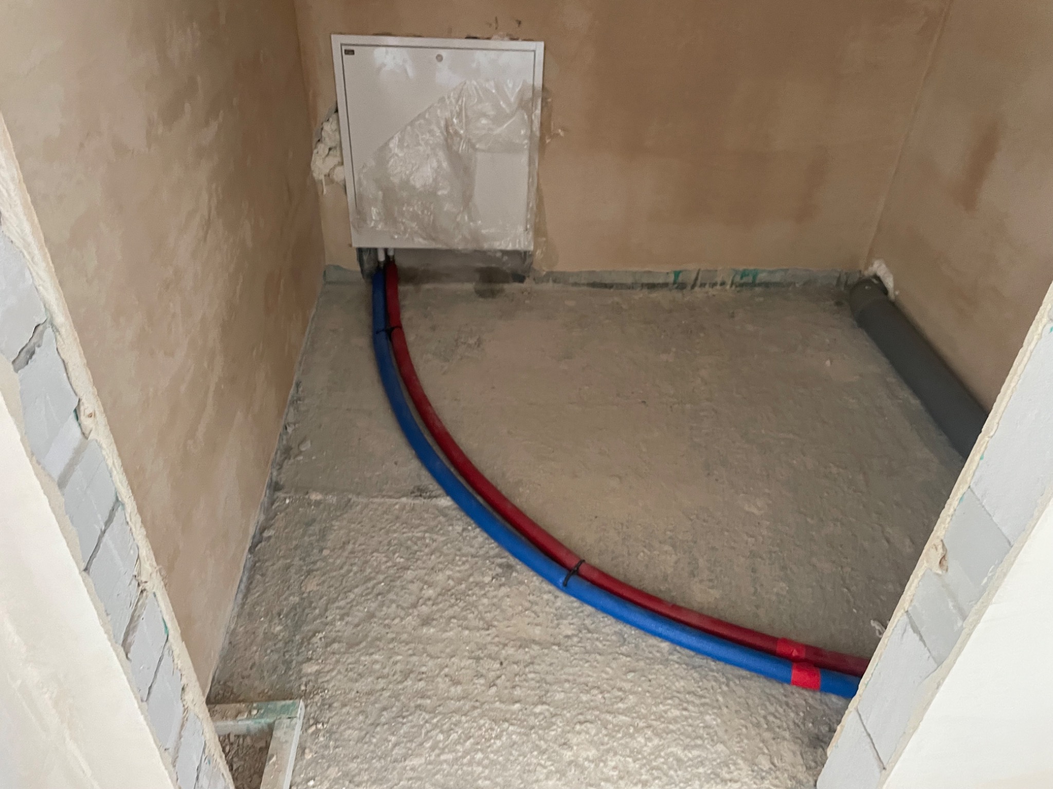 Surowa podłoga z wylewką w trakcie prac instalacyjnych, widoczne rury w kolorze niebieskim i czerwonym, zabezpieczona szafka instalacyjna na ścianie, surowe ściany w trakcie wykończenia.