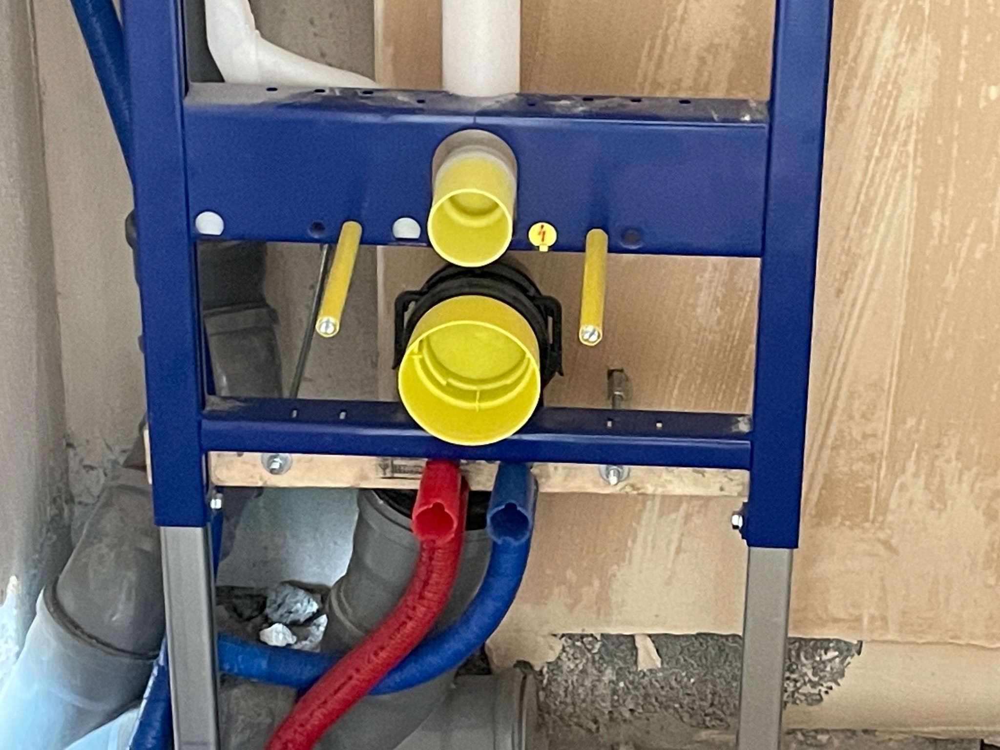 Stelaż podtynkowy WC z widocznymi przyłączami wody (czerwony i niebieski) oraz rurą odpływową, zamontowany w surowym stanie wykończenia łazienki.