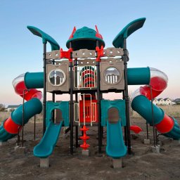 Big Toys Elżbieta Zygaryn - Nowoczesny plac zabaw z turkusowo-czerwonymi zjeżdżalniami otwartymi i tunelowymi, w tym jedną przezroczystą, na świeżo przygotowanym terenie z domami w tle.