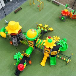 Big Toys Elżbieta Zygaryn - Widok z góry na rozbudowany, kolorowy zestaw do zabawy dla dzieci z tunelami, zjeżdżalniami, huśtawkami i elementami dekoracyjnymi, na zielonej nawierzchni bezpiecznej, obok budynku.