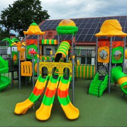 Big Toys Elżbieta Zygaryn - Nowoczesny, wieloelementowy zestaw zabawowy w żywych kolorach zieleni, żółci i pomarańczy, na zielonej nawierzchni bezpiecznej. W tle budynek z panelami fotowoltaicznymi.