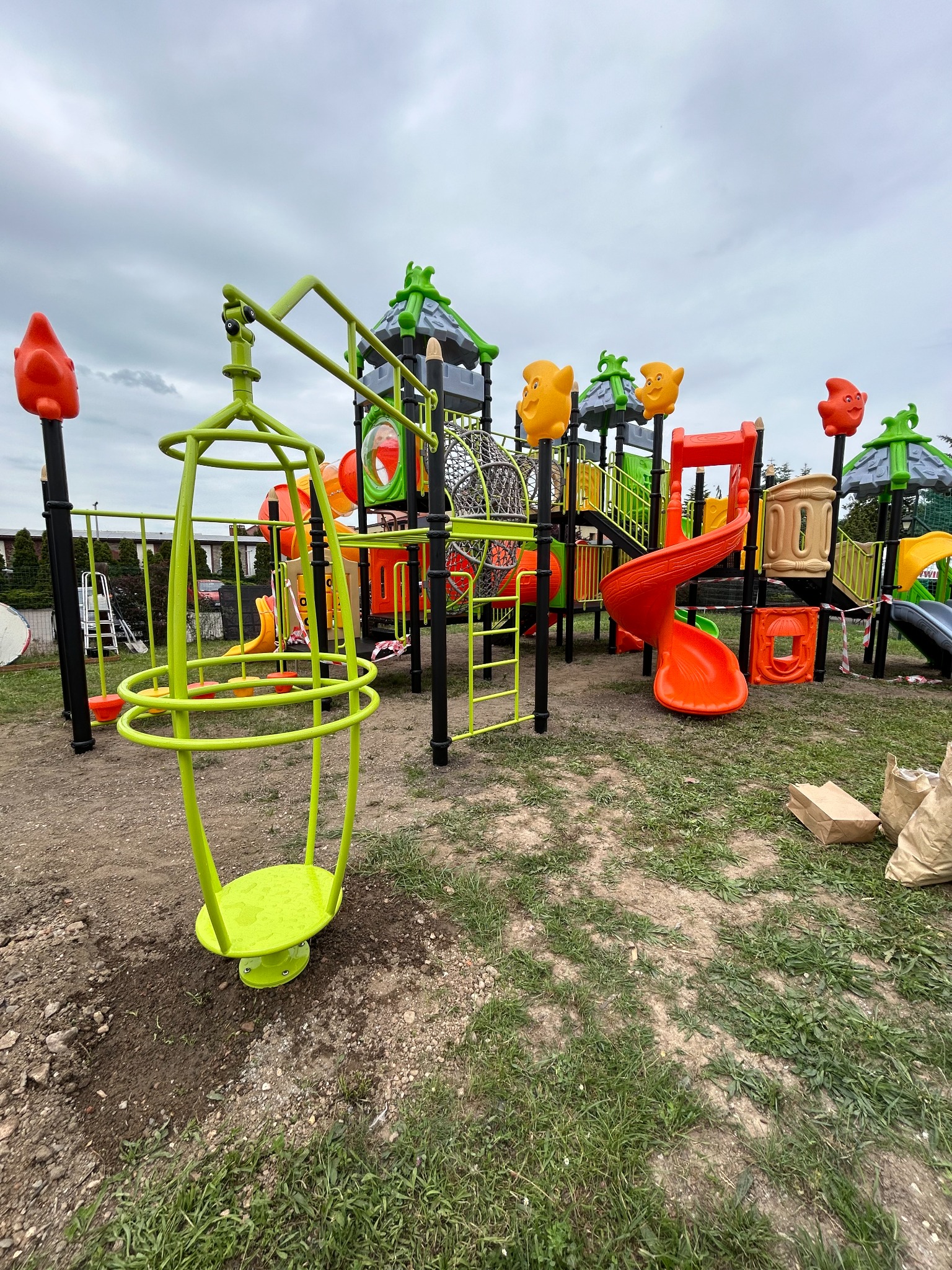 Kolorowy kompleks zabawowy z zieloną karuzelą obrotową na pierwszym planie, widoczne worki z piaskiem na ziemi, pochmurne niebo w tle.