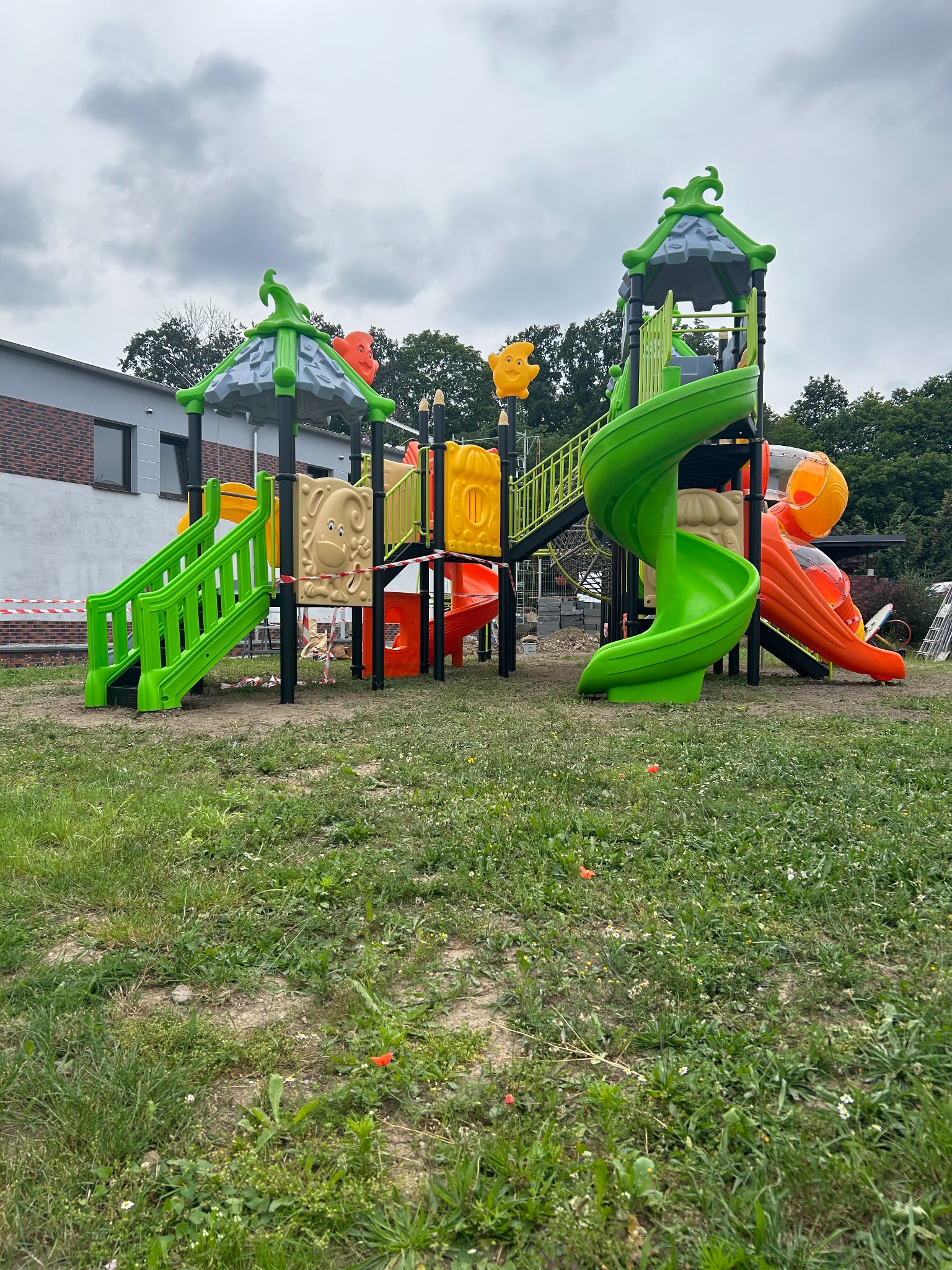 Kolorowy zestaw zabawowy z zieloną spiralną zjeżdżalnią i pomarańczową zjeżdżalnią tunelową, otoczony trawą i częściowo ogrodzony taśmą ostrzegawczą.