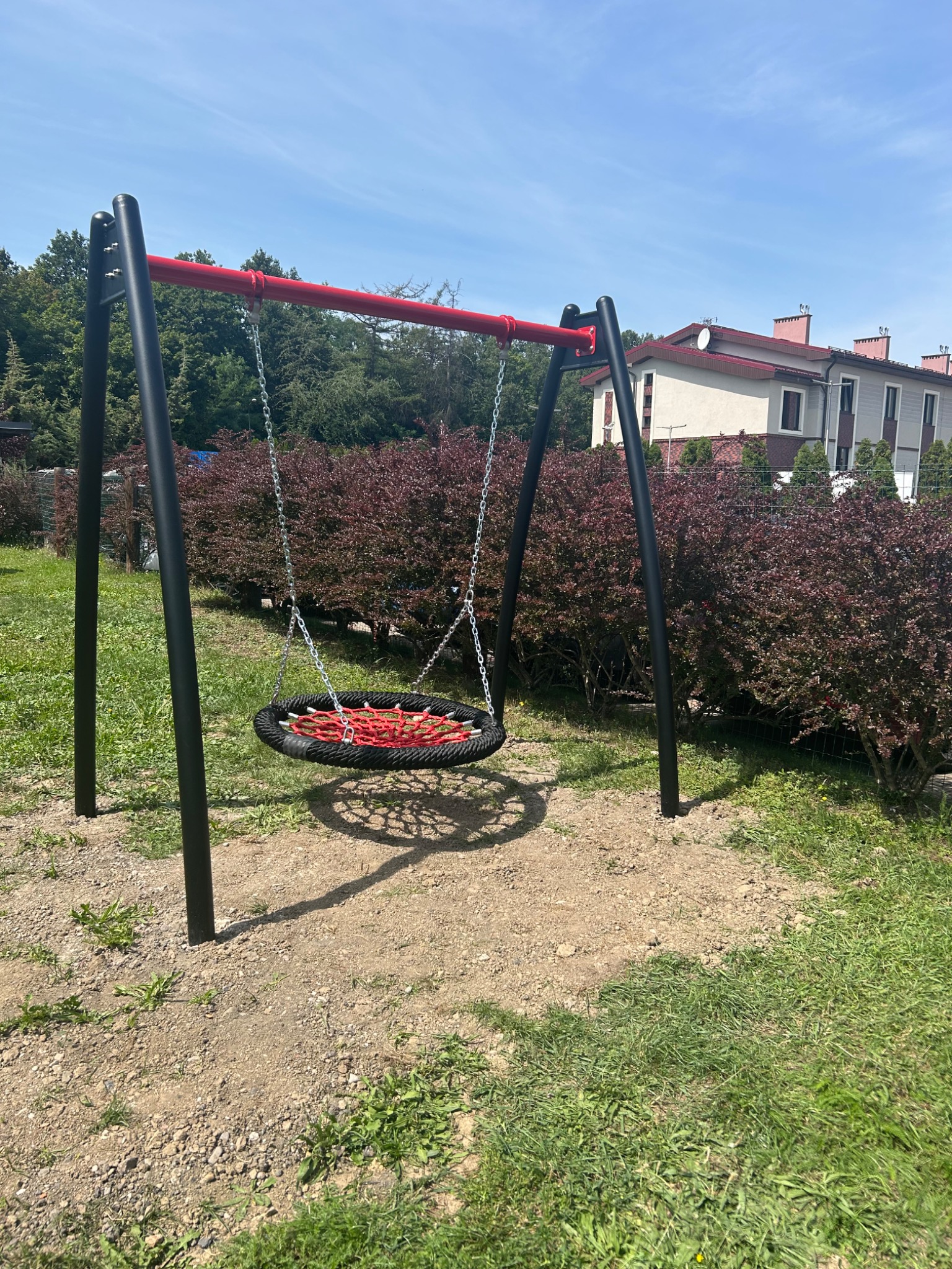 Huśtawka typu bocianie gniazdo z czarną ramą i czerwonym siedziskiem na tle budynku mieszkalnego i krzewów.