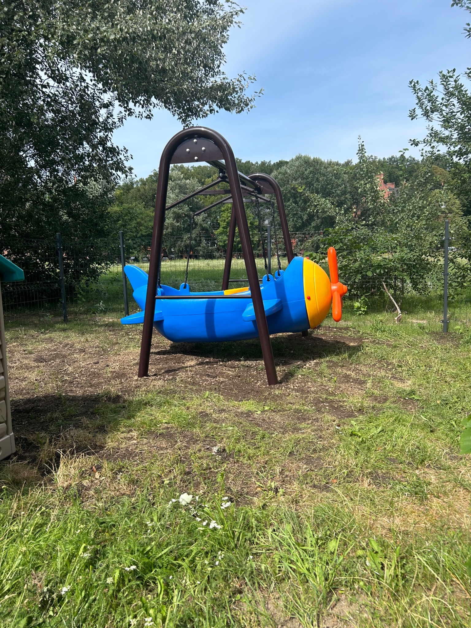 Huśtawka w kształcie niebieskiego samolotu z żółto-pomarańczowym śmigłem na placu zabaw, otoczona zielenią i drzewami.