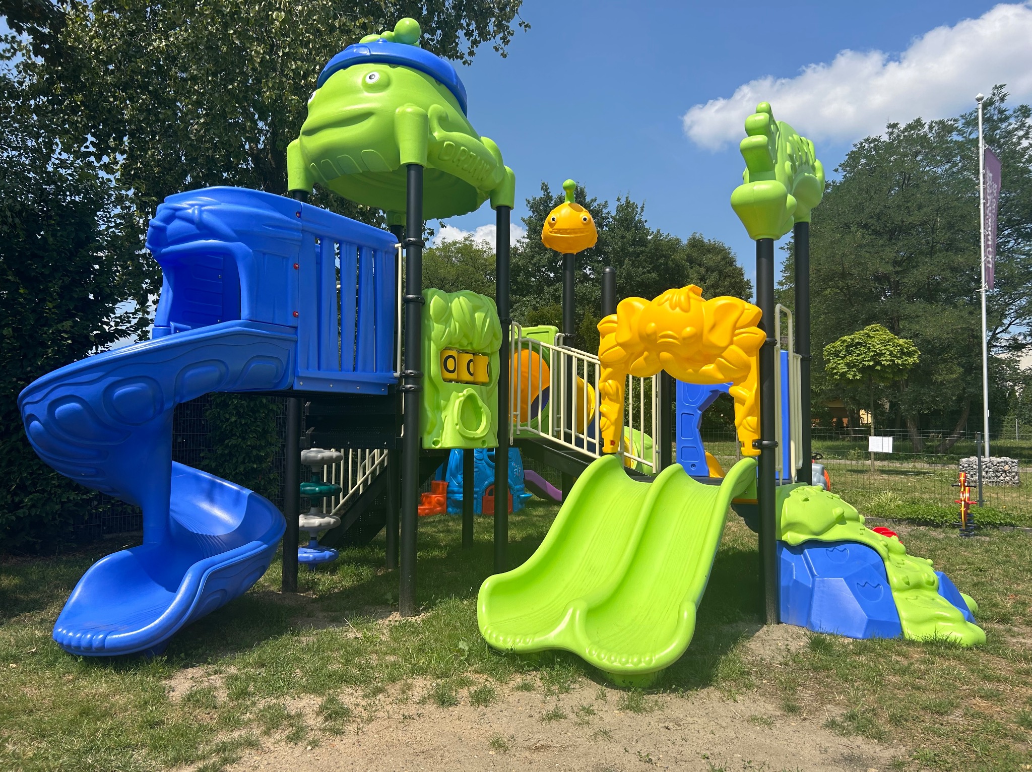 Kolorowy zestaw zabawowy z dwiema zjeżdżalniami, tunelem i elementami w kształcie zwierząt, umieszczony na trawiastym terenie z drzewami w tle.