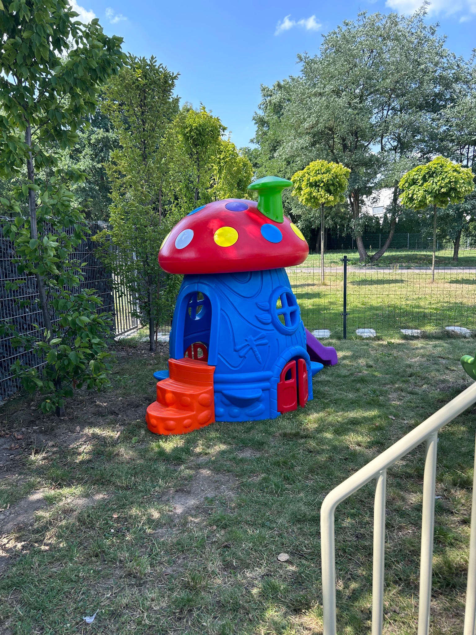 Kolorowy domek dla dzieci w kształcie muchomora z pomarańczowymi schodkami i małą zjeżdżalnią, umieszczony na trawniku w ogrodzie, otoczony drzewami i siatką ogrodzeniową.