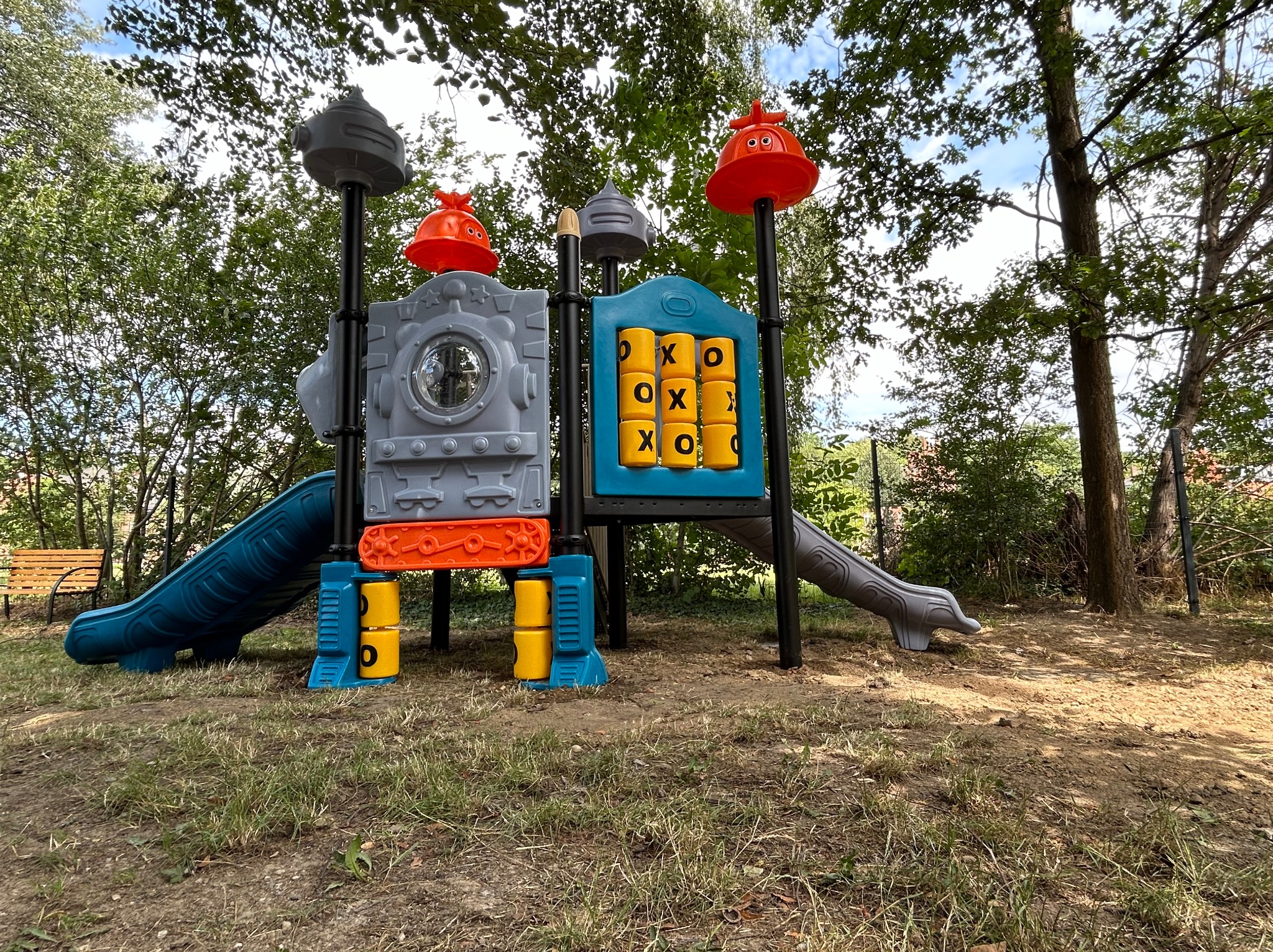 Kolorowy zestaw zabaw dla dzieci z dwiema zjeżdżalniami i panelem do gry w kółko i krzyżyk, umieszczony na trawiastym terenie pod drzewami.