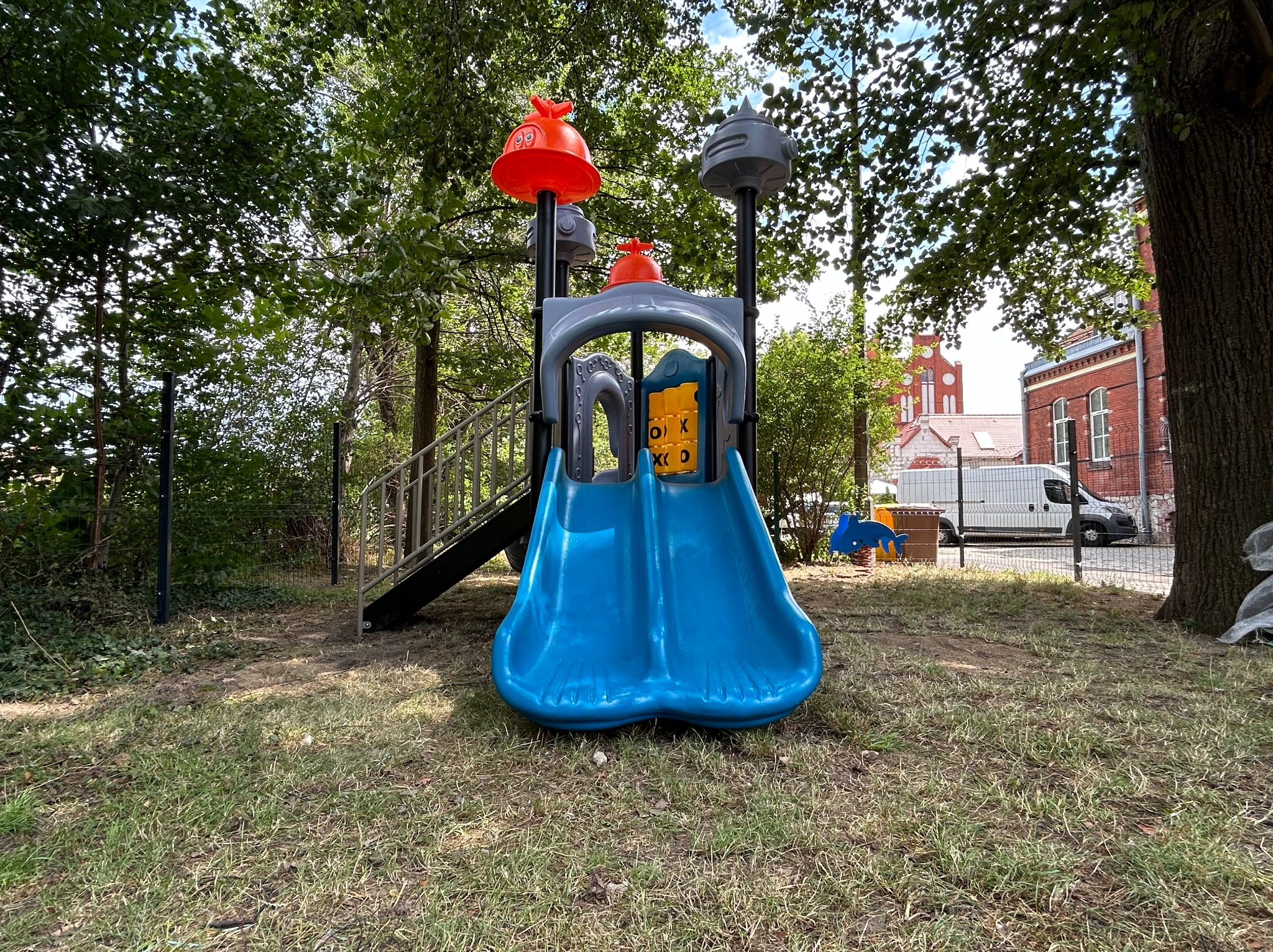 Kolorowy plac zabaw z niebieską zjeżdżalnią, elementami w kształcie statków kosmicznych i grą OX w tle, otoczony zielenią z widocznym budynkiem z czerwonej cegły.