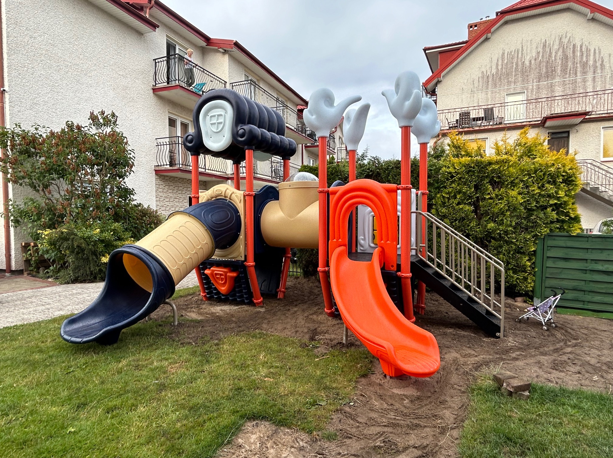 Kolorowy zestaw zabawowy z dwiema zjeżdżalniami i elementami w kształcie rąk, umieszczony na trawniku przed budynkami mieszkalnymi.