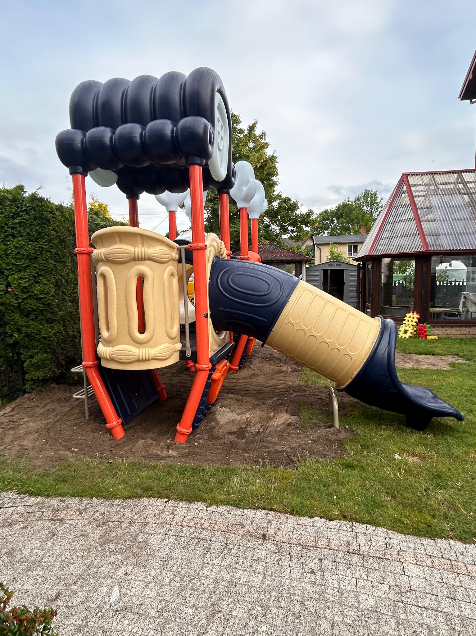 Kolorowy zestaw zabaw dla dzieci w kształcie pojazdu z beżową zjeżdżalnią tunelową i oponami na szczycie, ustawiony na trawie obok domu z oranżerią.