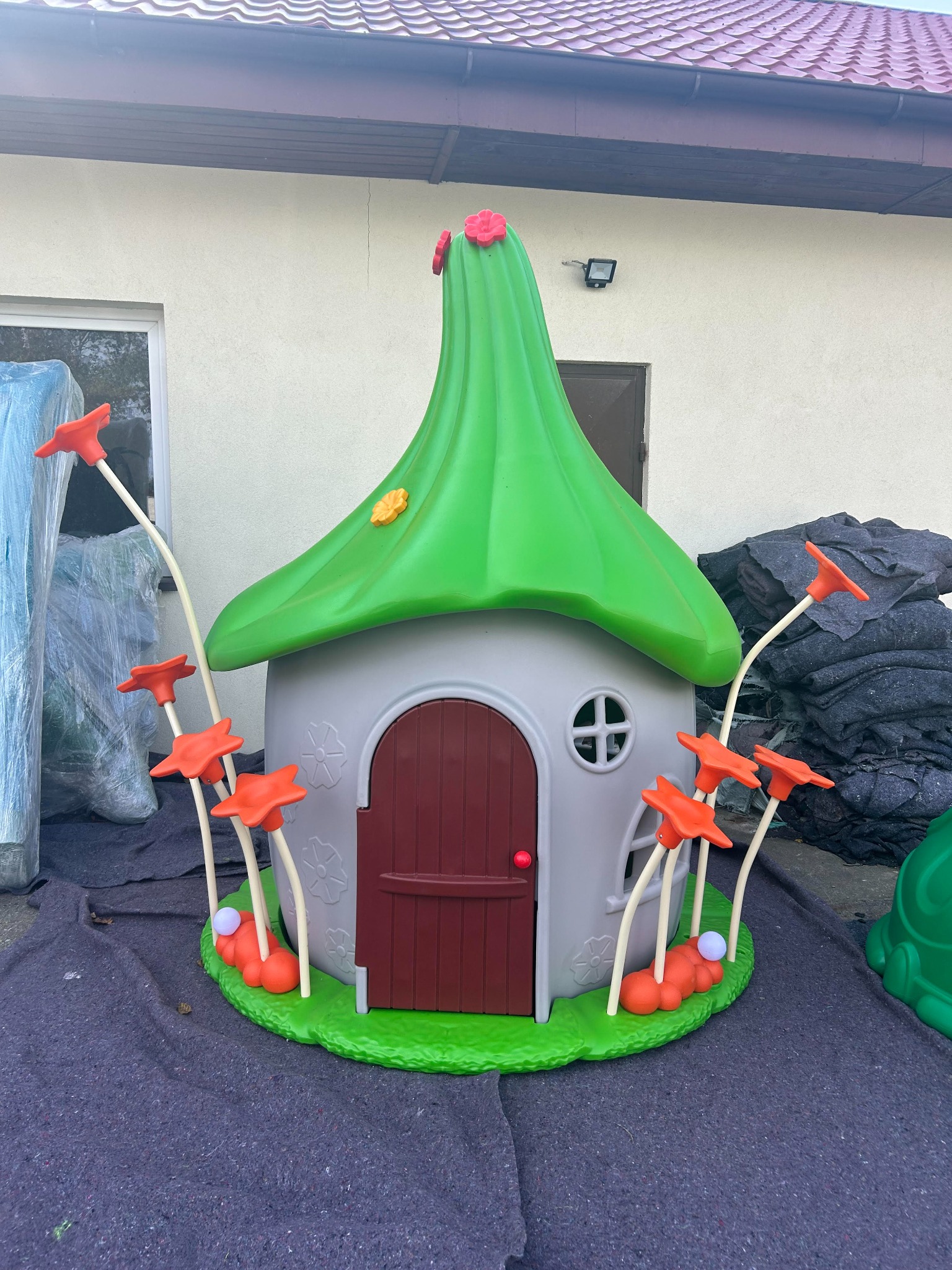 Plastikowy domek dla dzieci w kształcie grzyba z brązowymi drzwiami i pomarańczowymi kwiatami na łodygach, stojący na zielonej podstawie imitującej trawę.