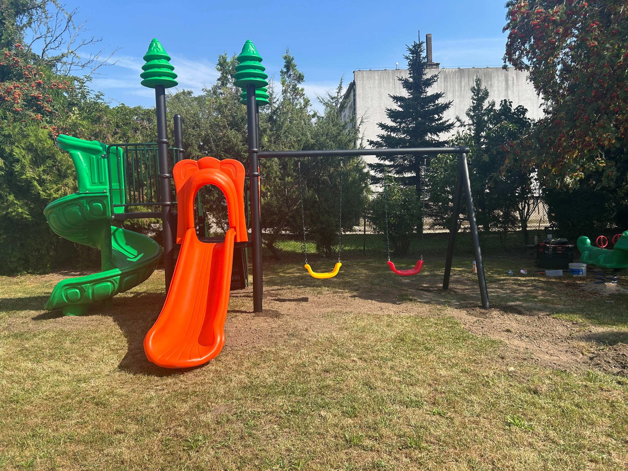 Kolorowy zestaw zabawowy z zieloną zjeżdżalnią spiralną, pomarańczową prostą zjeżdżalnią i huśtawką z żółtym i czerwonym siedziskiem na trawiastym terenie.