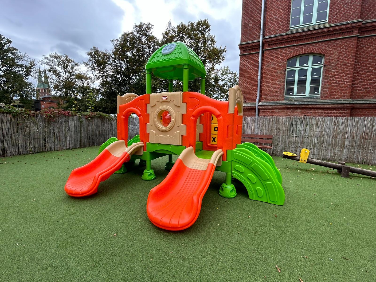 Kolorowy zestaw zabaw dla dzieci z dwiema zjeżdżalniami i wieżą, na zielonej sztucznej trawie, na tle ceglanego budynku. Widoczna huśtawka wagowa.