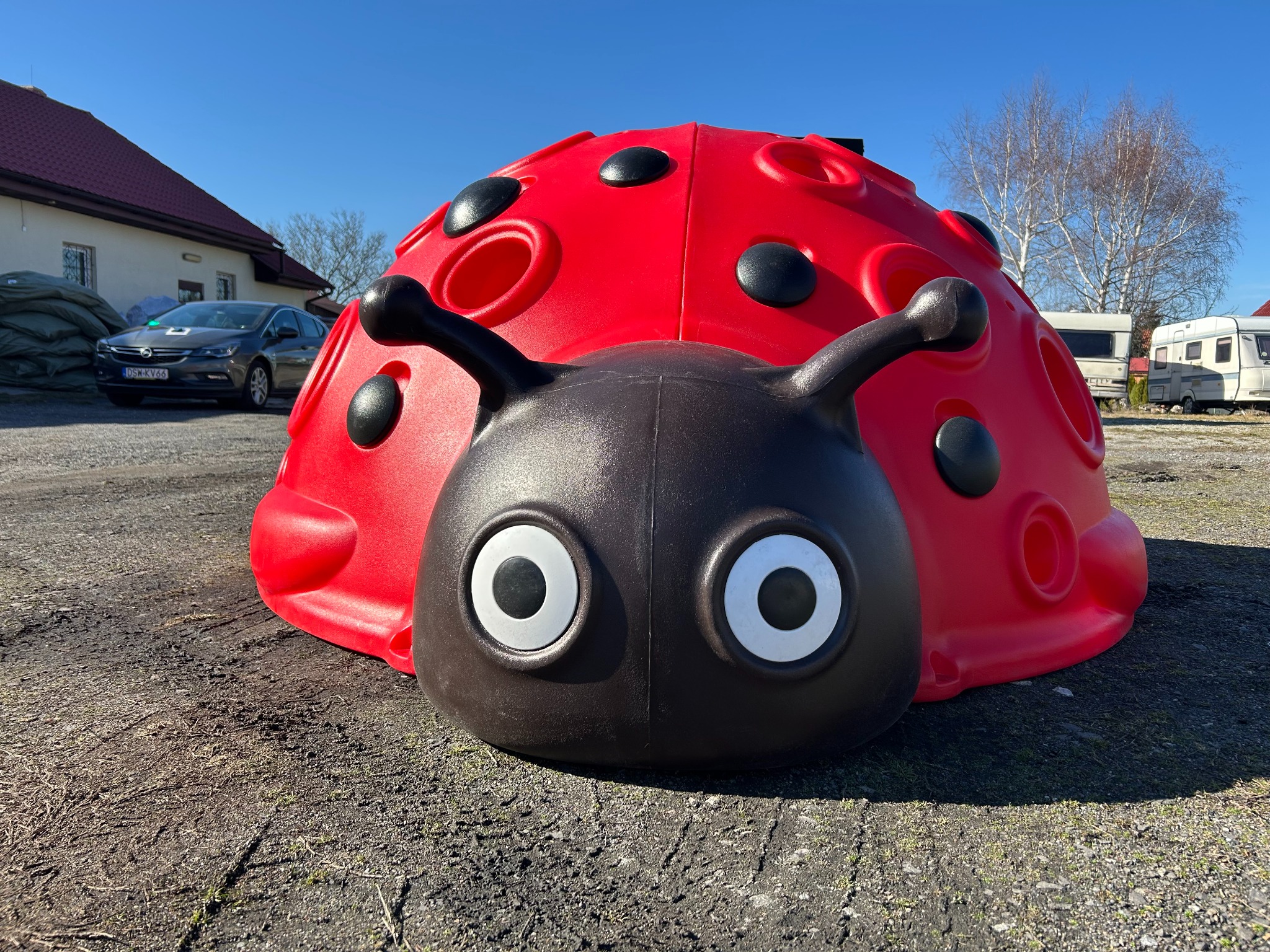 Kopuła wspinaczkowa w kształcie biedronki, czerwono-czarna, z dużymi oczami, na żwirowym podłożu, w tle budynek i przyczepy kempingowe, w słoneczny dzień.