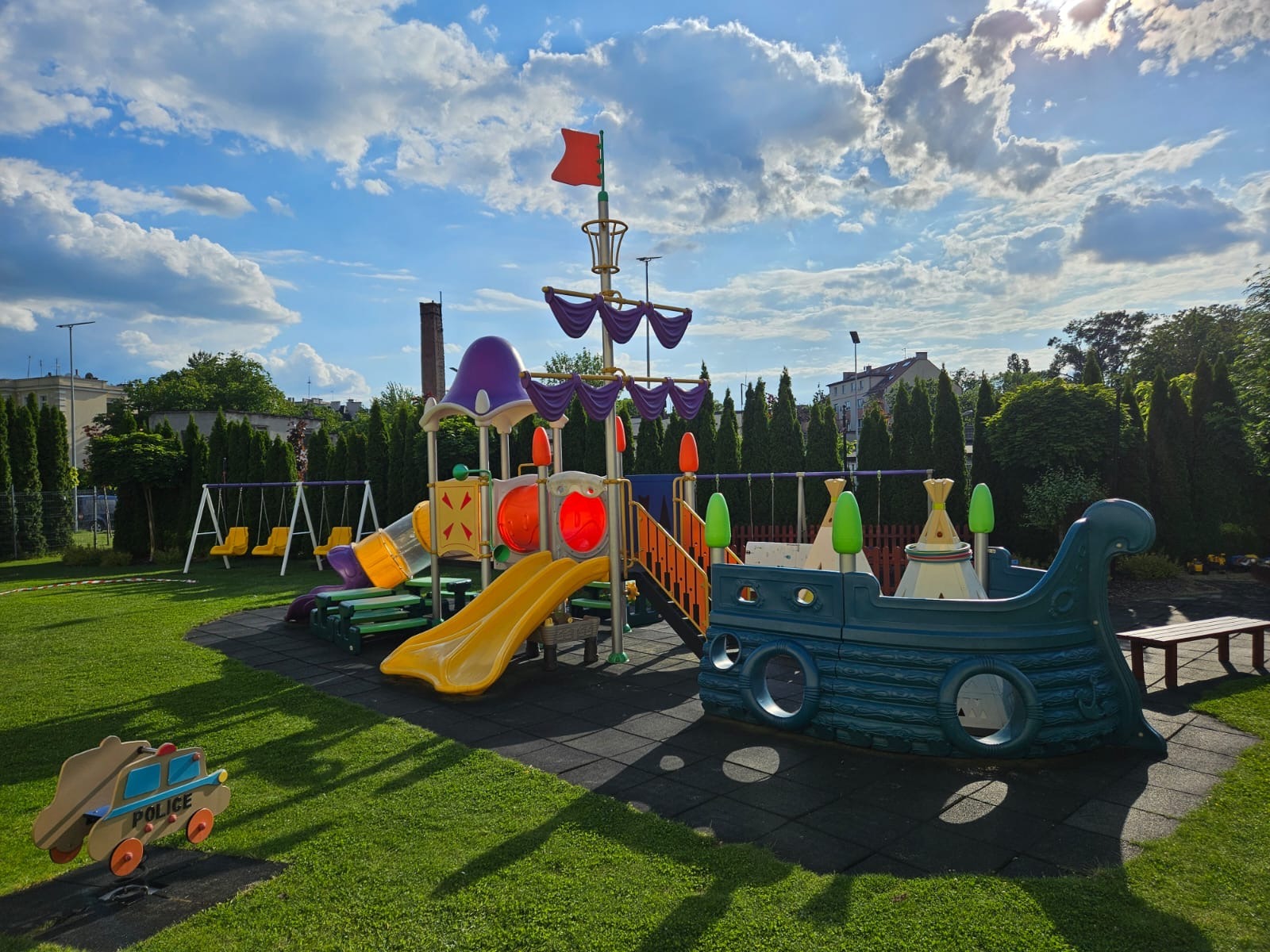 Kolorowy plac zabaw z motywem statku pirackiego i huśtawkami na tle zielonego trawnika i błękitnego nieba z chmurami. W tle budynki i drzewa.