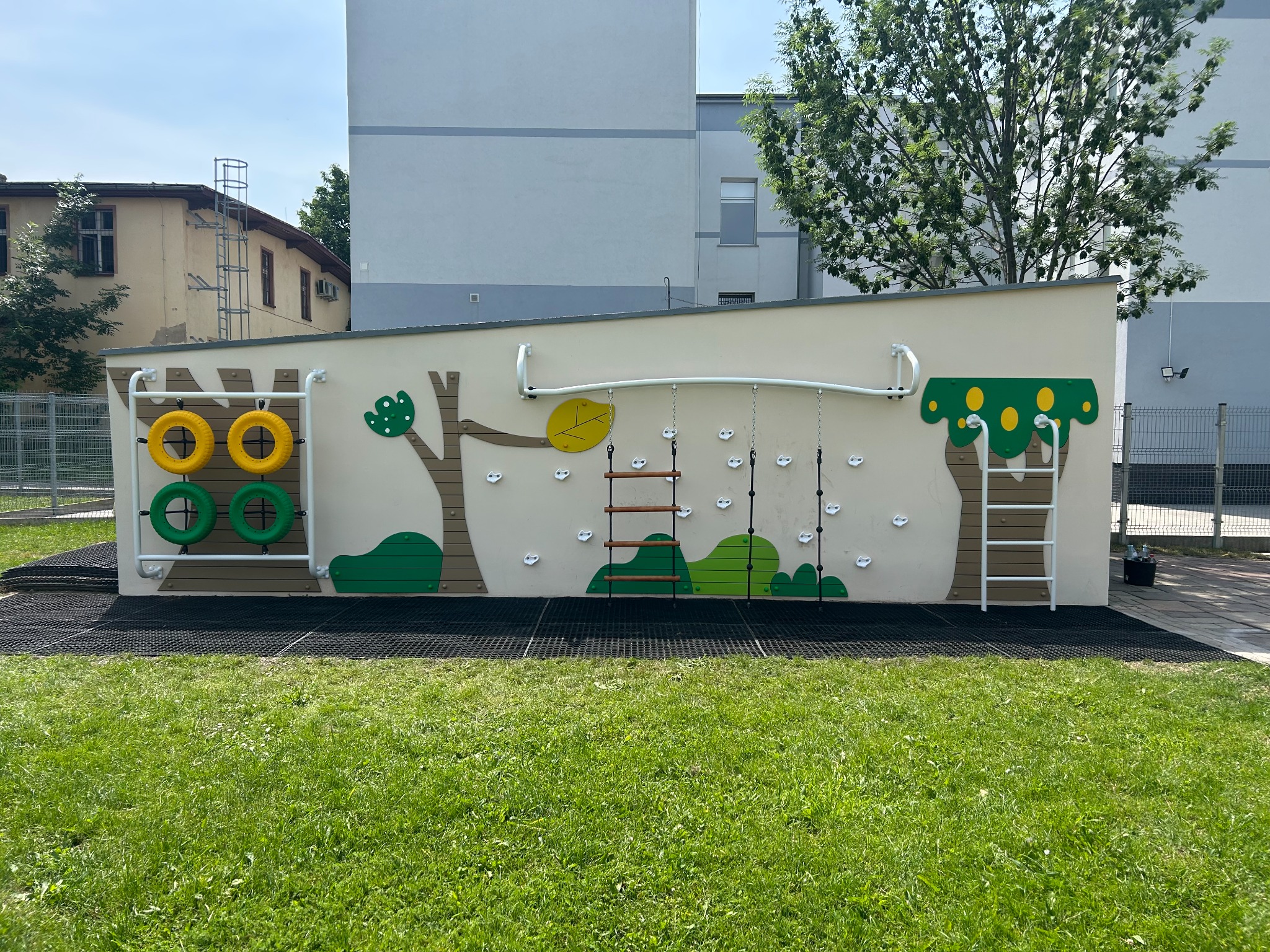 Kolorowy plac zabaw z elementami wspinaczkowymi i drabinkami na tle zielonego trawnika i budynku. Elementy dekoracyjne w formie drzew.