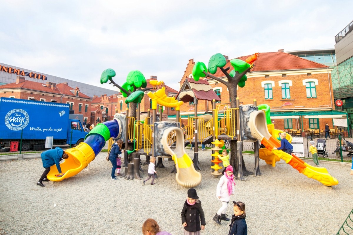 Kolorowy plac zabaw z motywem drzew, zjeżdżalniami i dziećmi bawiącymi się na żwirowym podłożu, na tle budynków miejskich i nieba.