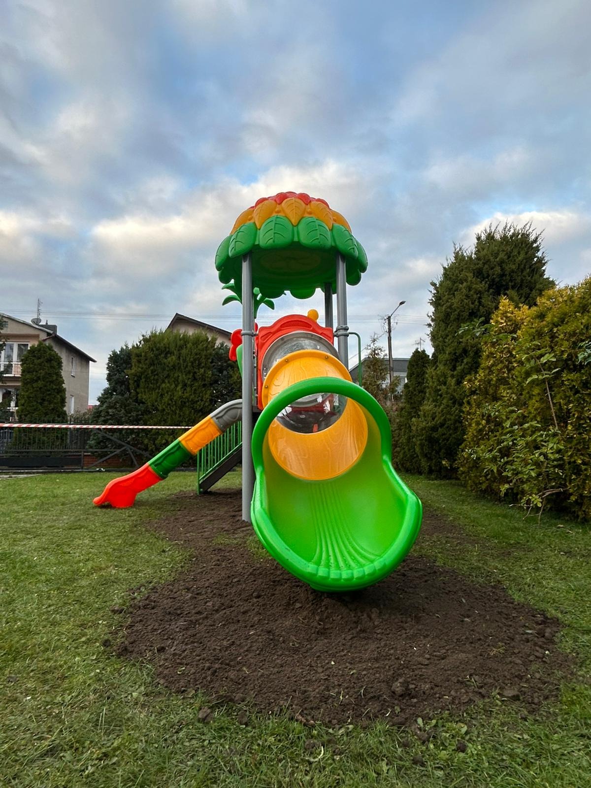 Kolorowa zjeżdżalnia z tunelem na trawniku, otoczona zielenią. Element placu zabaw w kształcie lokomotywy z motywem kwiatowym na szczycie.