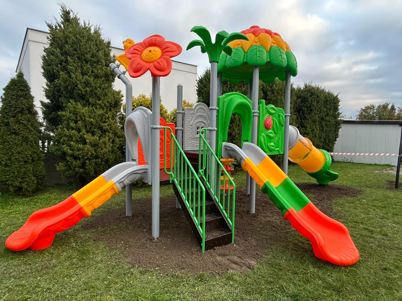 Kolorowy plac zabaw z dwiema zjeżdżalniami, schodkami i elementami dekoracyjnymi w kształcie kwiatów i drzew, otoczony zielonym trawnikiem i krzewami.