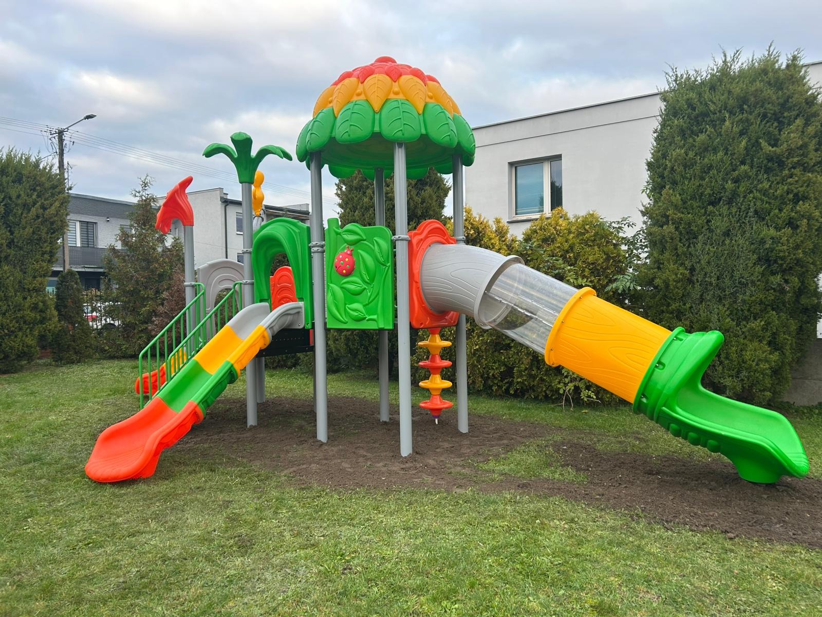 Kolorowy plac zabaw z dwiema zjeżdżalniami, tunelem i motywem biedronki, ustawiony na trawniku przed budynkiem mieszkalnym w pochmurny dzień.