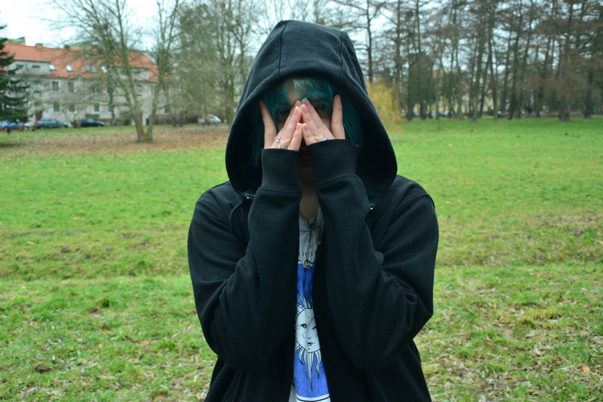 Osoba w czarnej bluzie z kapturem, zasłaniająca twarz dłońmi z pierścionkiem, na tle zielonego trawnika i drzew w parku.