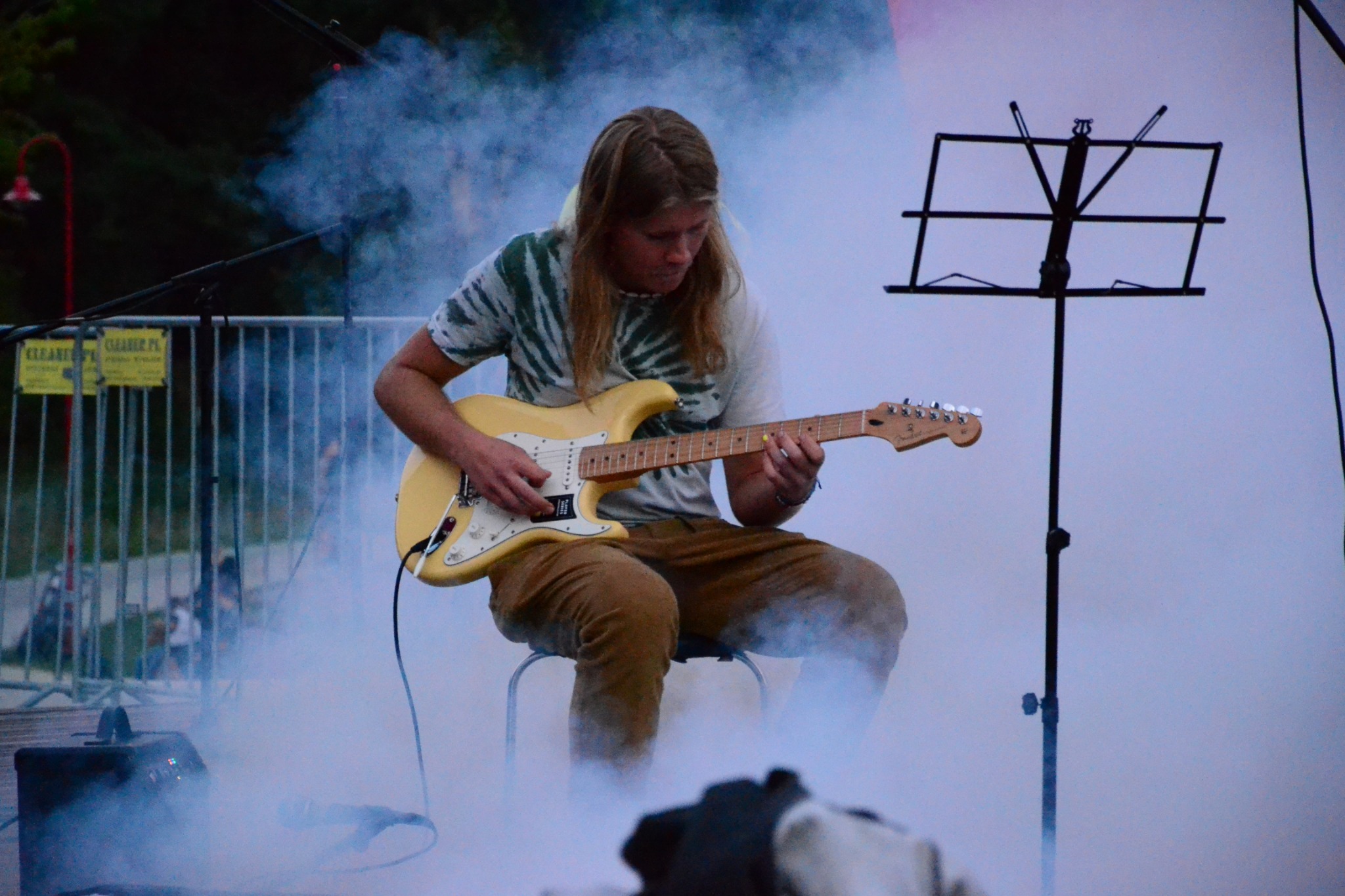 Młody mężczyzna z długimi włosami gra na jasnej gitarze elektrycznej siedząc na krześle, otoczony dymem, z nutami na statywie w tle.