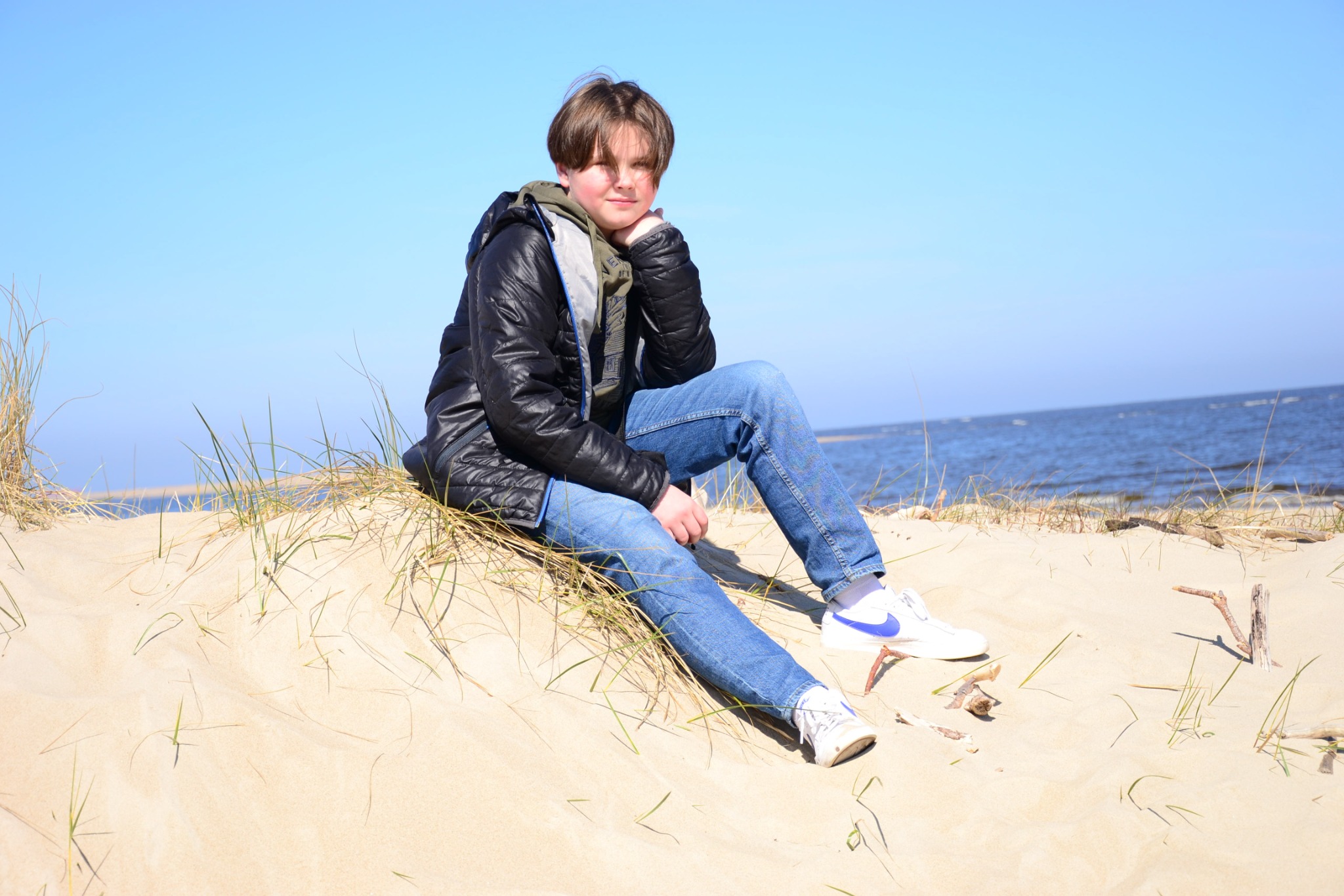 Młody mężczyzna w czarnej kurtce i jeansach siedzi na piaszczystej wydmie z widokiem na morze w słoneczny dzień.