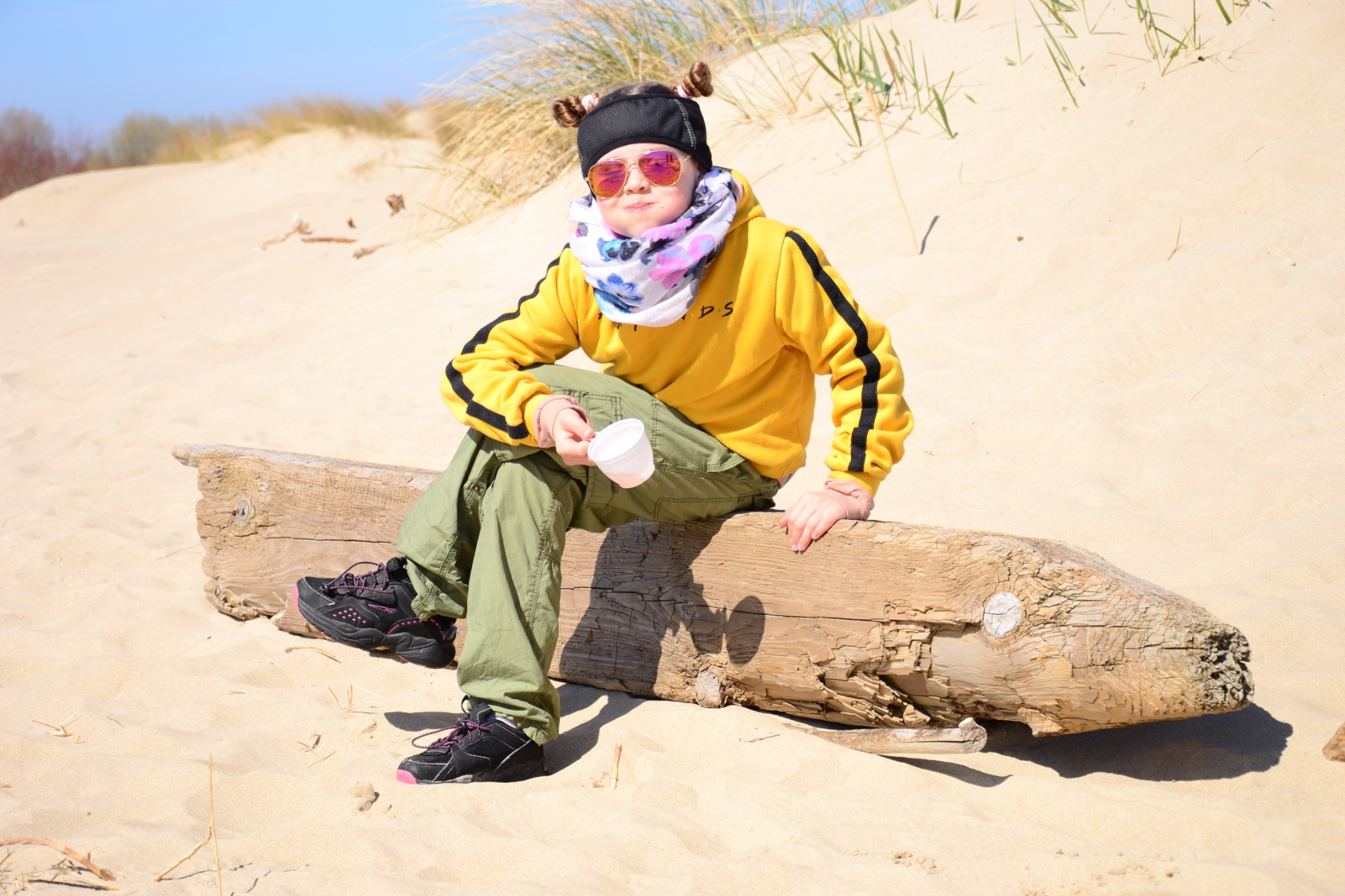 Dziewczynka w żółtej bluzie i okularach przeciwsłonecznych siedzi na spróchniałym pniu na piaszczystej plaży, trzymając w ręku biały kubek.