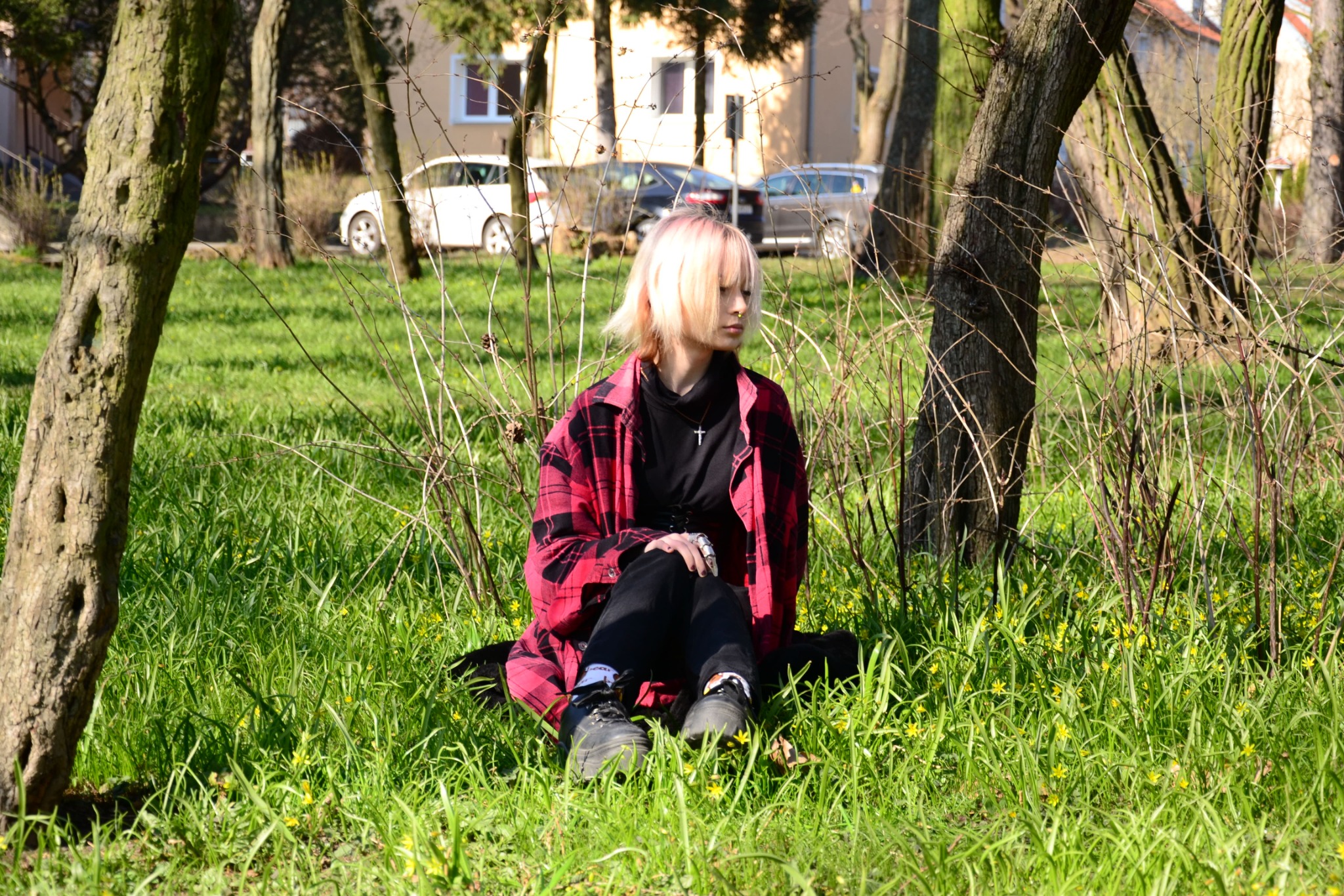 Młoda kobieta z blond włosami siedzi na trawie w parku, ubrana w czarne spodnie, czarną bluzkę i czerwoną koszulę w kratę, z widocznym naszyjnikiem z krzyżykiem, otoczona drzewami i zieloną trawą...