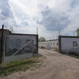 Budowa parterowego budynku z otwartą bramą wjazdową, widoczne pręty zbrojeniowe, stos materiałów budowlanych i ślady opon na piaszczystym podłożu.