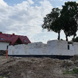 Wznoszenie ścian parterowego domu z białych bloczków, widoczne zbrojenie fundamentów i częściowo wykończony dach sąsiedniego budynku z czerwoną dachówką.