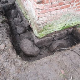 Firma Guzowski - Odkopany fundament ceglanego budynku z widocznymi kamieniami, ziemią i narzędziem do kopania.