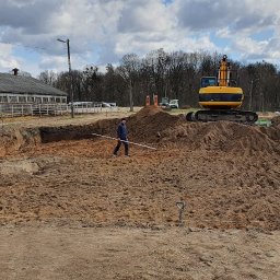 Firma Guzowski - Wykop pod fundament domu z widoczną koparką i pracownikiem z poziomicą, na tle zabudowania gospodarczego i pochmurnego nieba.